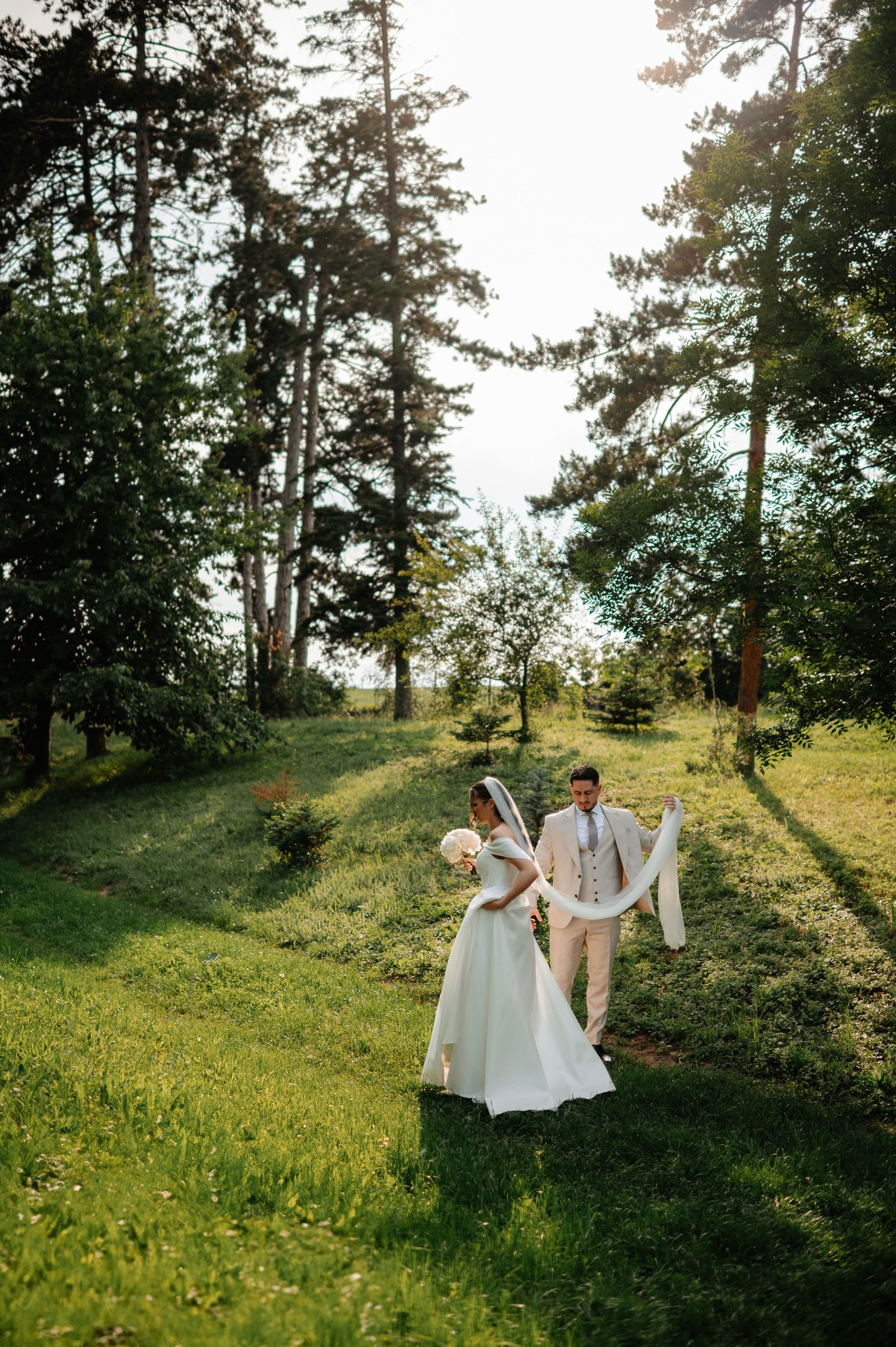 Bride and groom walking through sunlit forest clearing, groom carrying flowing white bridal veil.