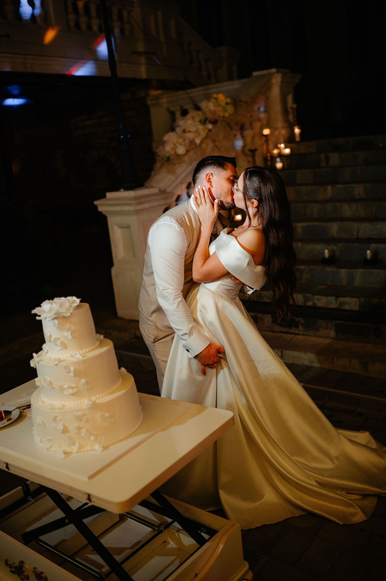 Bride and groom sharing a romantic kiss beside a white three-tier wedding cake at an elegant outdoor night reception.