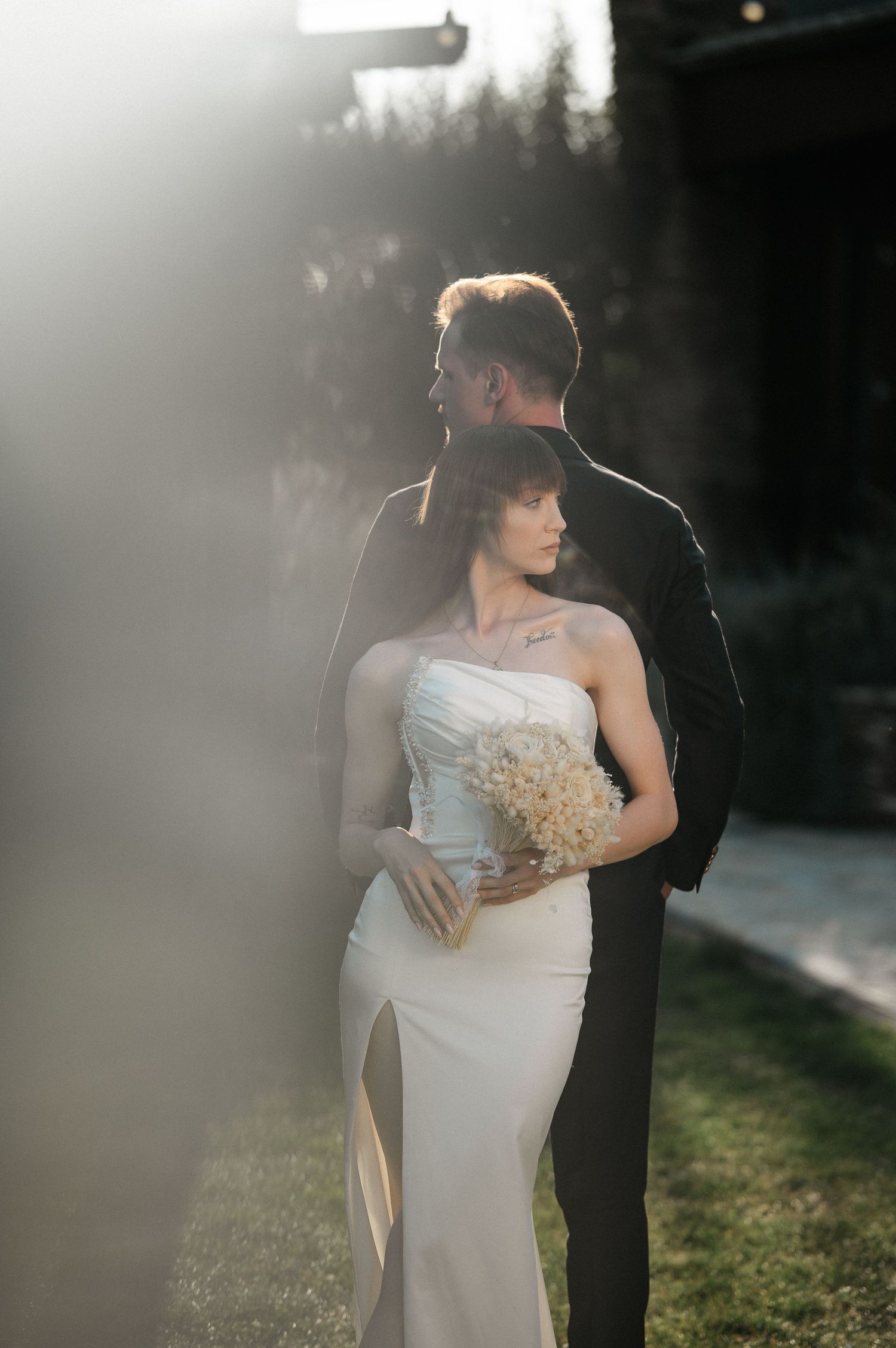 Groom embraces bride from behind as she holds bouquet in dreamy soft light wedding portrait.
