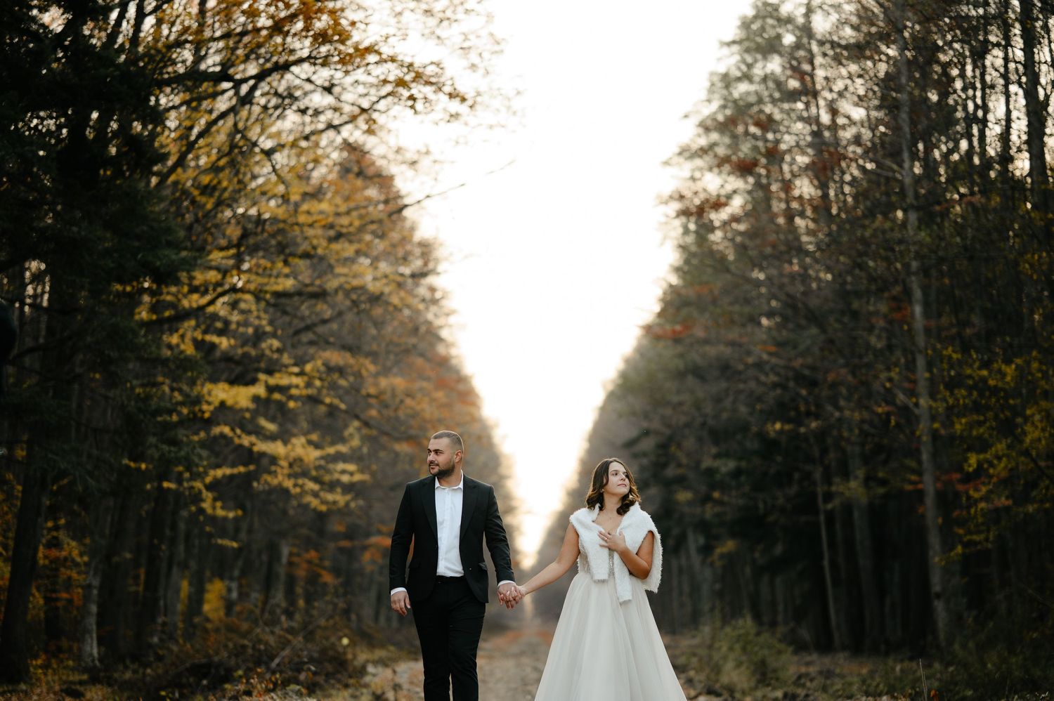 Bride and groom holding hands on a forest path surrounded by autumn foliage during an outdoor fall wedding.