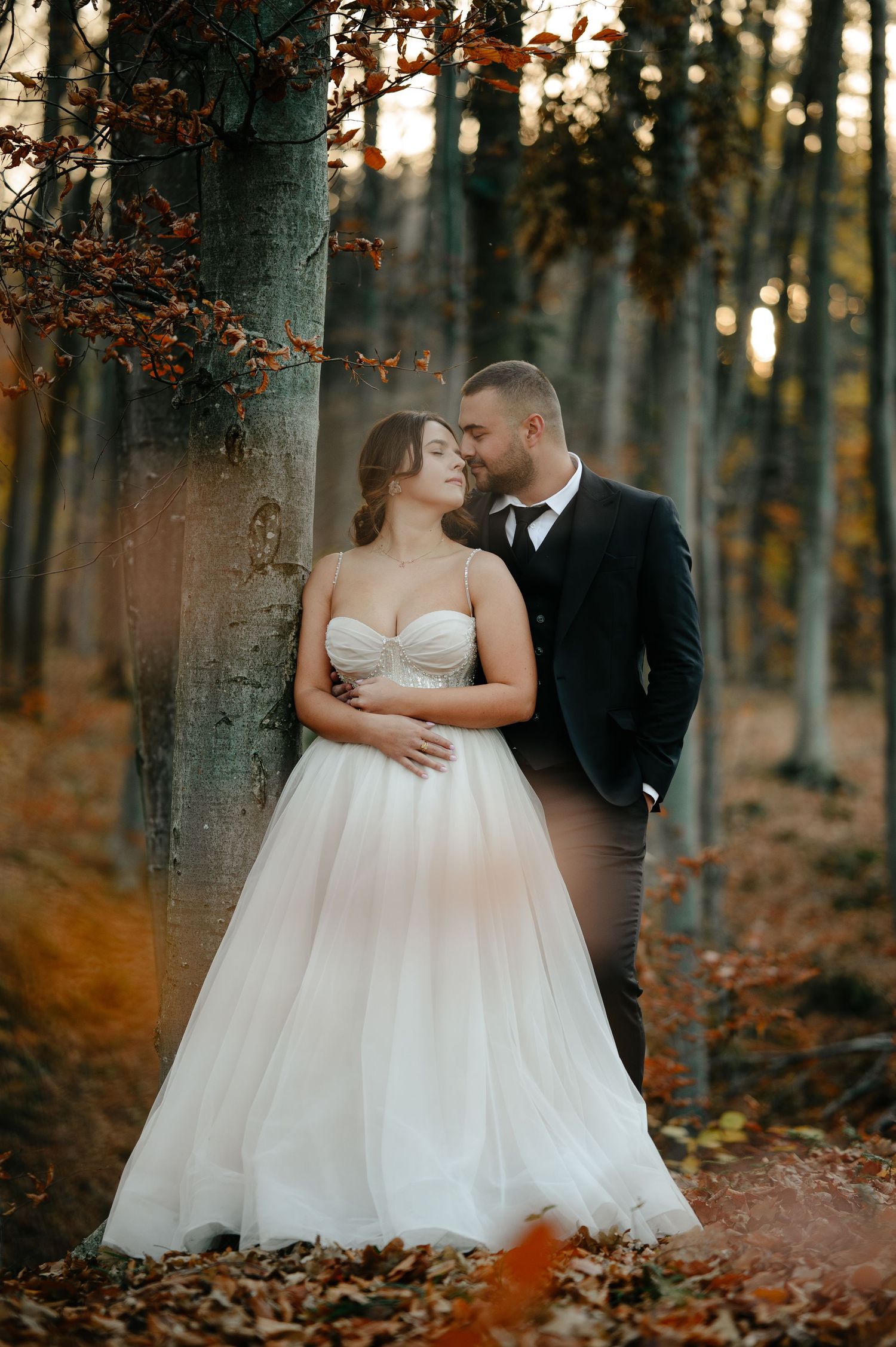 A bride and groom share a romantic moment in an autumn forest, surrounded by golden leaves and tall trees.