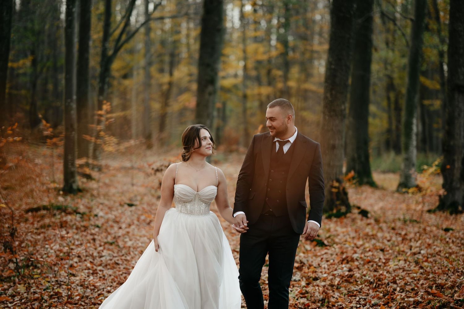 Bride and groom walking hand in hand through an autumn forest, surrounded by golden foliage and fallen leaves.