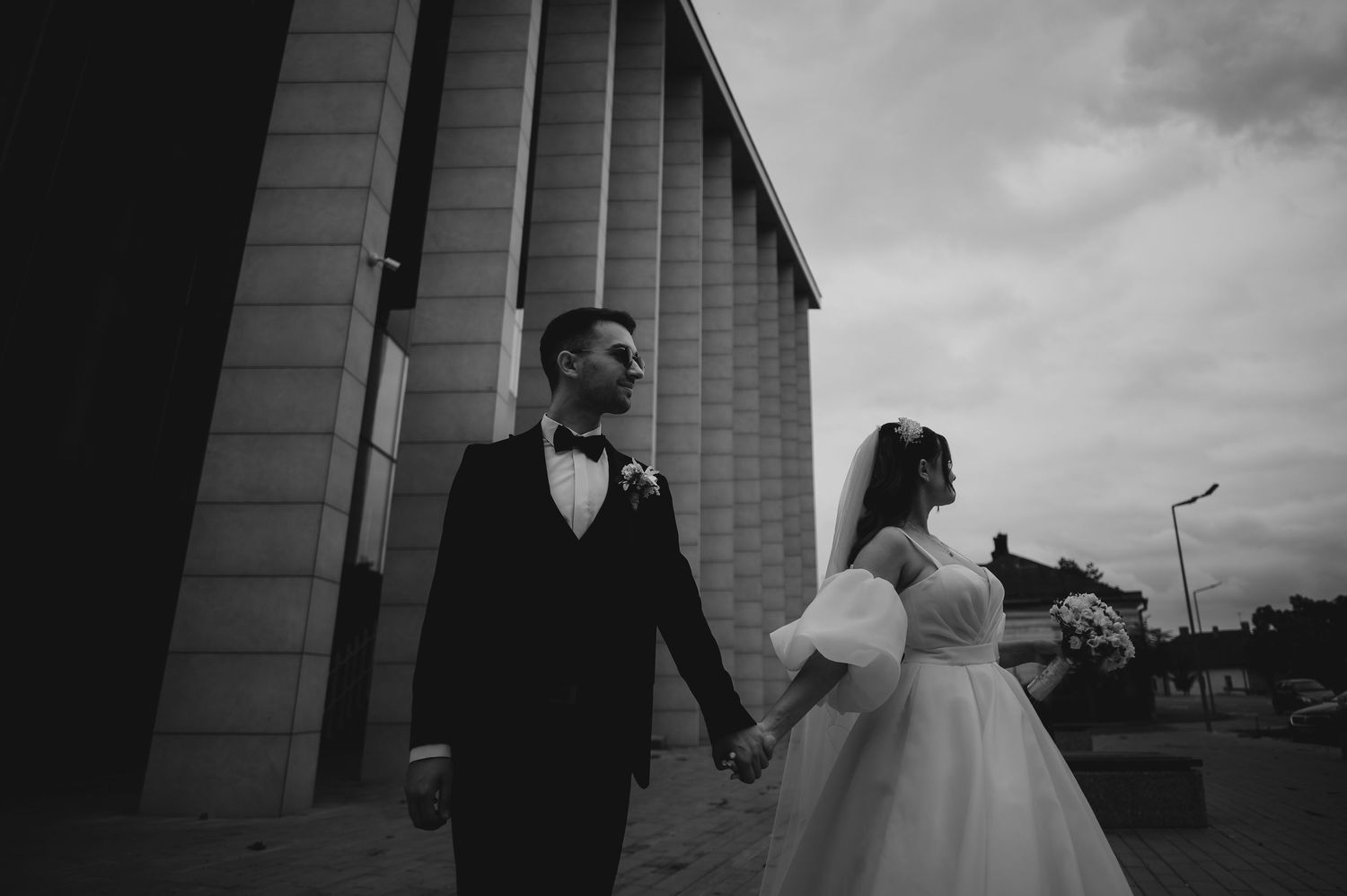 Black and white photo of bride and groom holding hands near building.