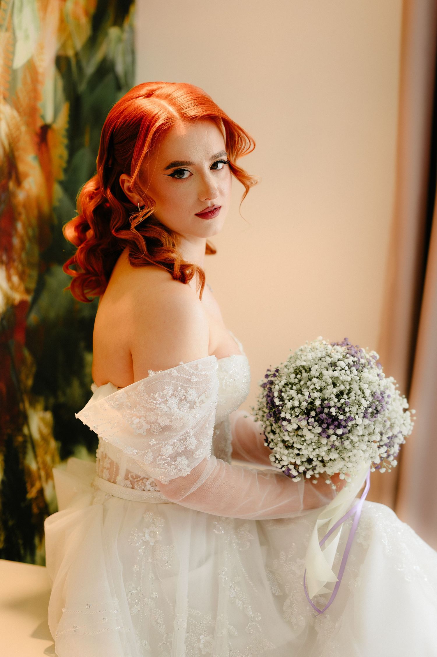 Red-haired bride in off-shoulder lace gown holds white and purple baby's breath bouquet, posing elegantly indoors.
