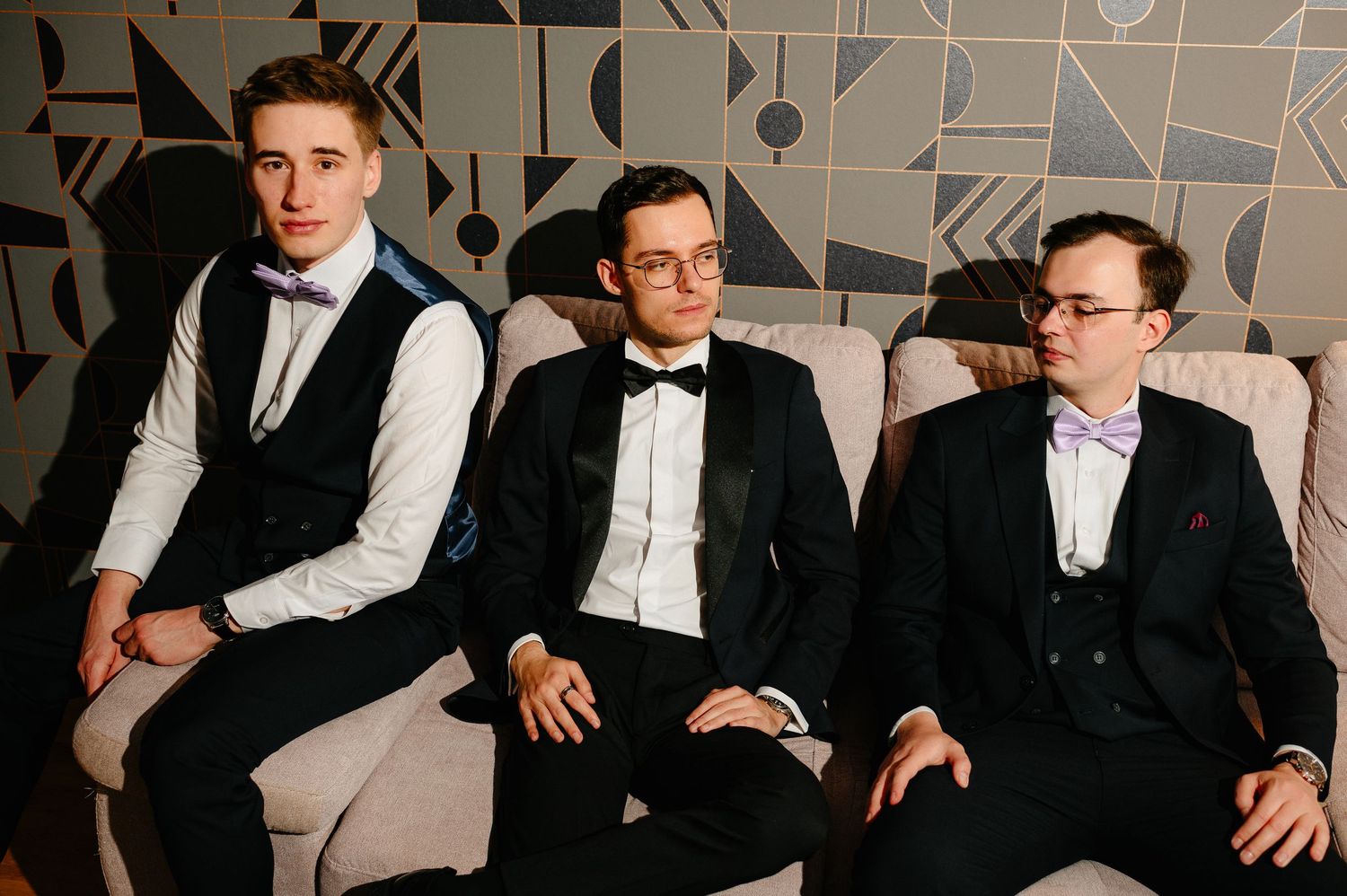 Three young men in formal tuxedos with bow ties sit together on a sofa against a geometric patterned wall.