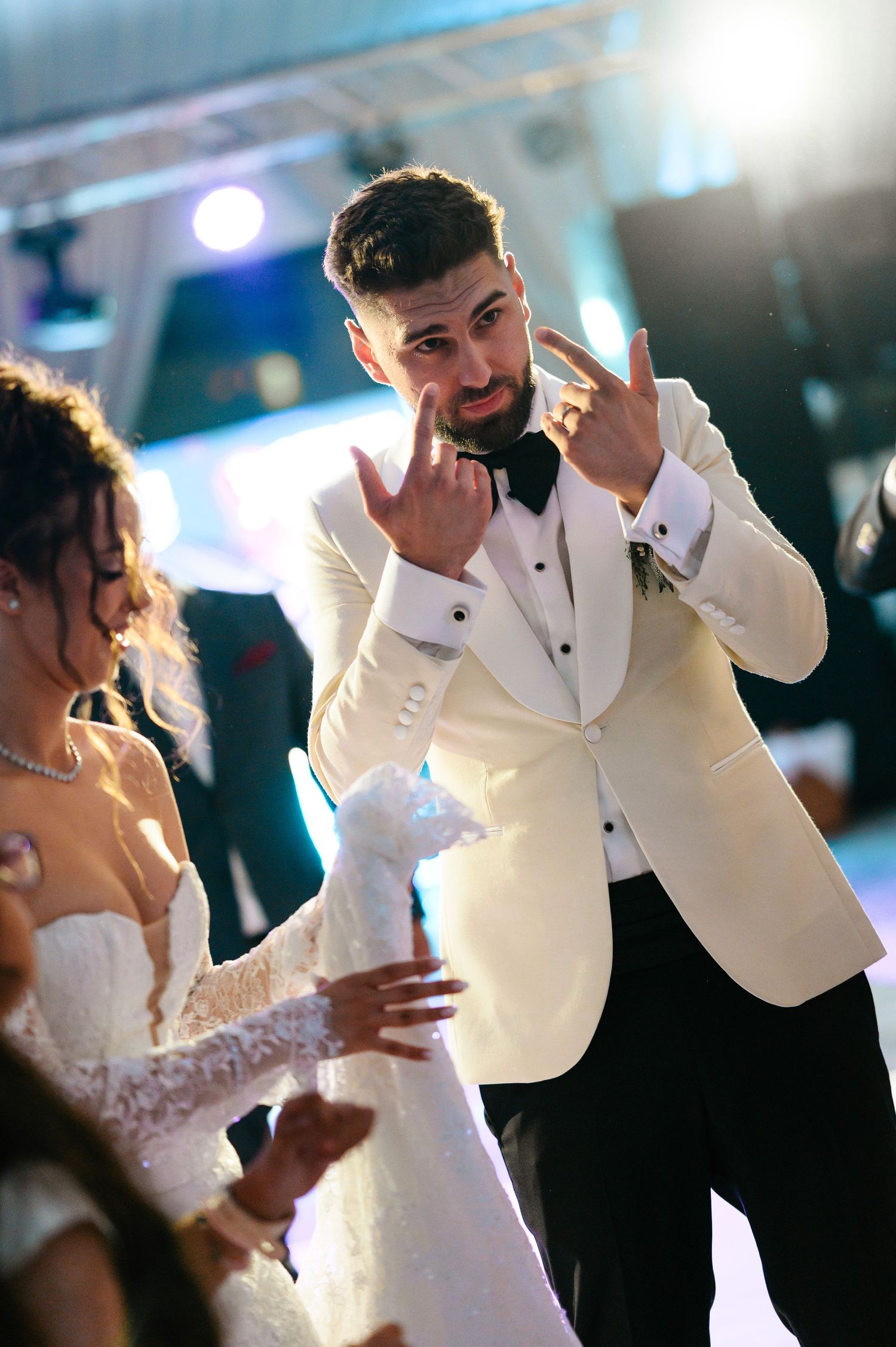 Groom in white tuxedo making rock hand gestures while dancing with bride in lace wedding dress at reception.