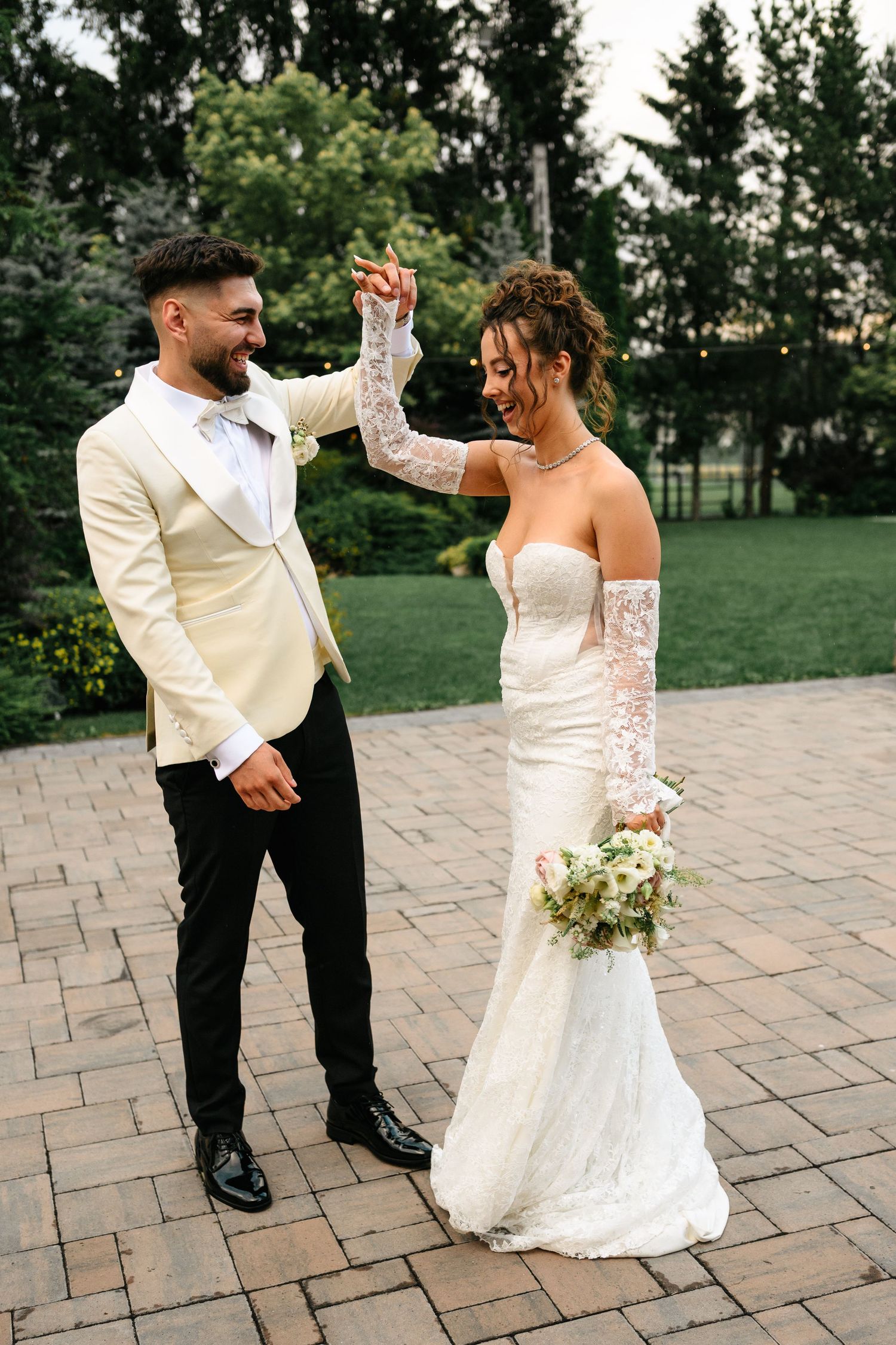 Bride and groom dancing outdoors, groom in ivory tuxedo, bride in lace wedding gown holding bouquet.