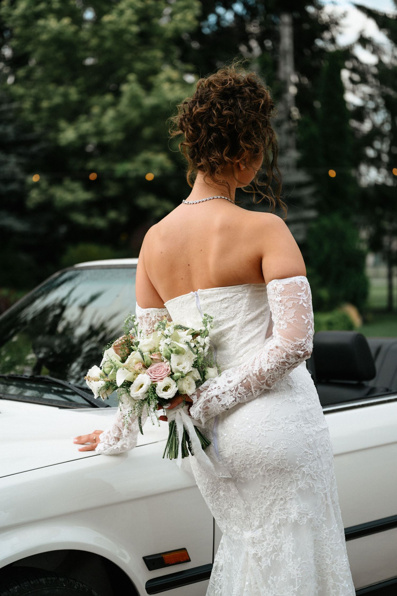 Bride in white lace strapless gown holding bouquet of white and pink flowers, standing beside a white convertible car.