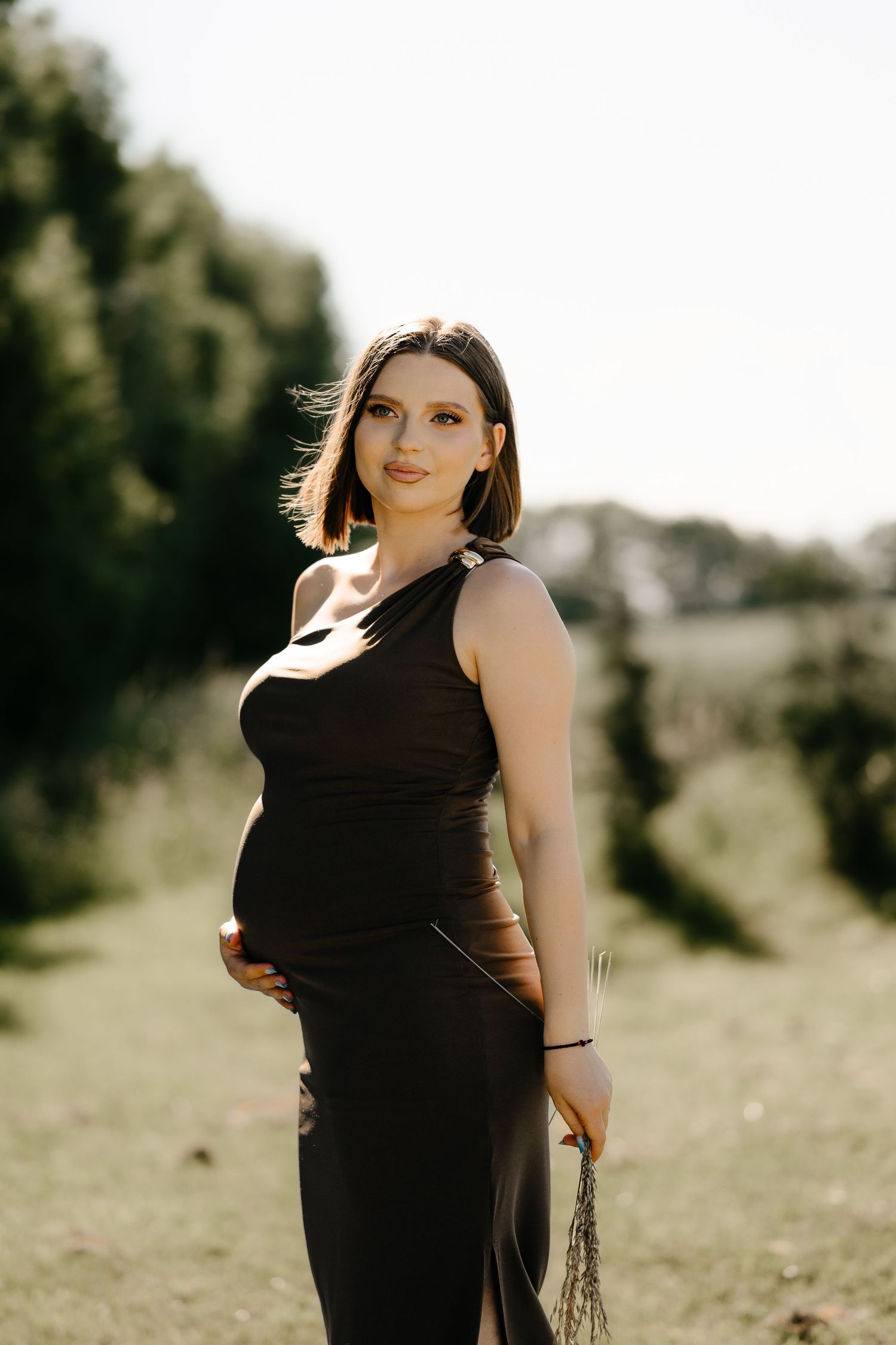 Pregnant woman in black one-shoulder dress holding wildflowers in sunny field.