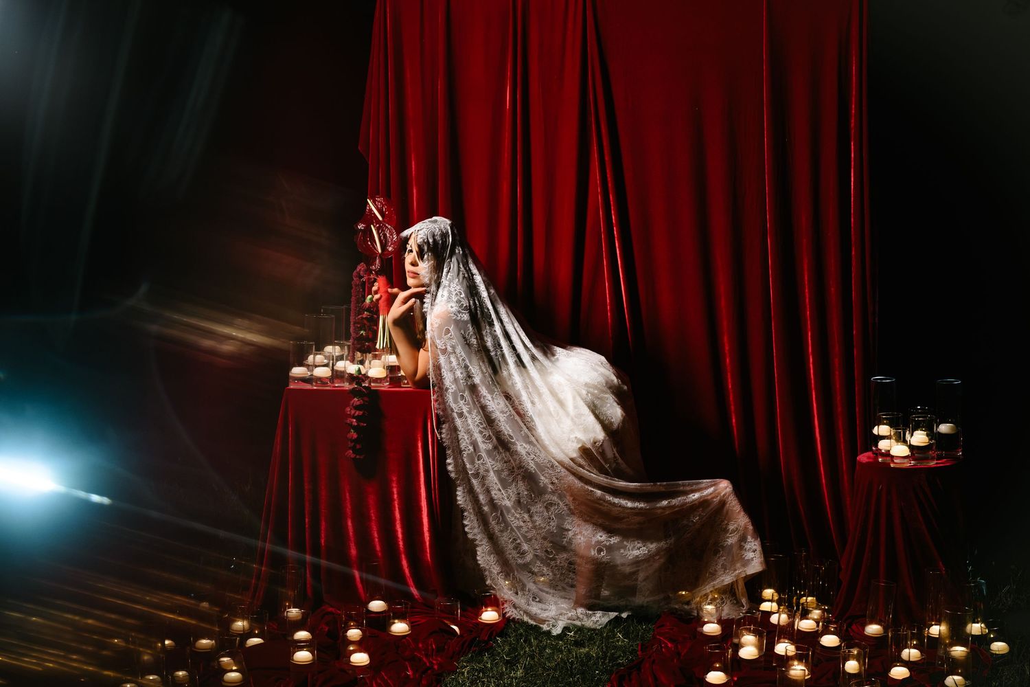 Bride in lace veil seated by red velvet curtains surrounded by glowing candles in dramatic dark wedding setting.