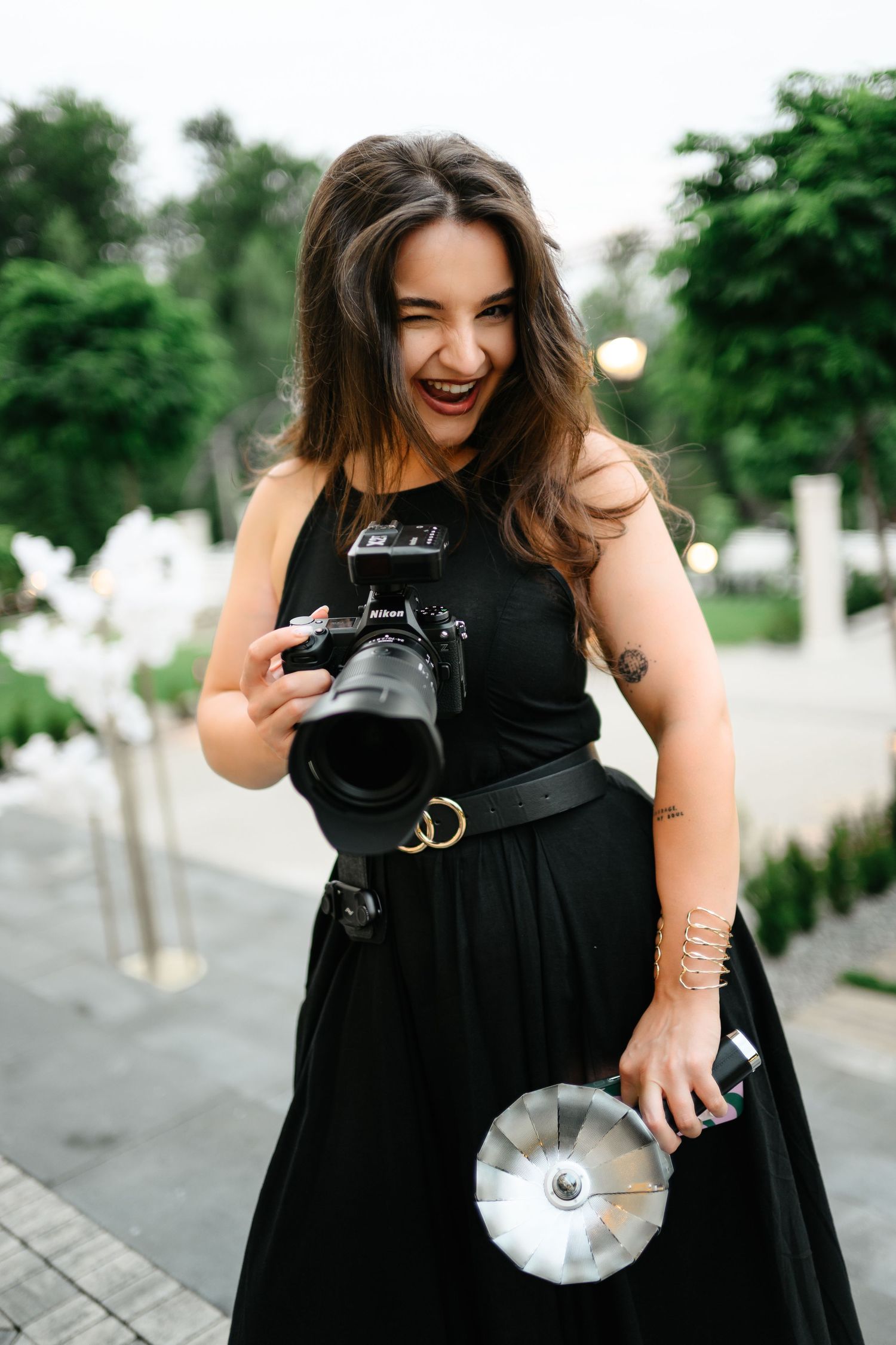 Smiling brunette woman in black dress holding a DSLR camera and flash reflector outdoors in a park.