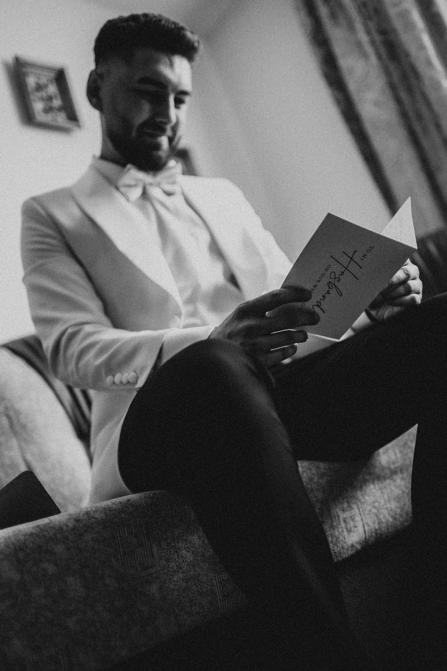 Groom in white tuxedo jacket and bow tie reads a heartfelt card while seated, captured in black and white.