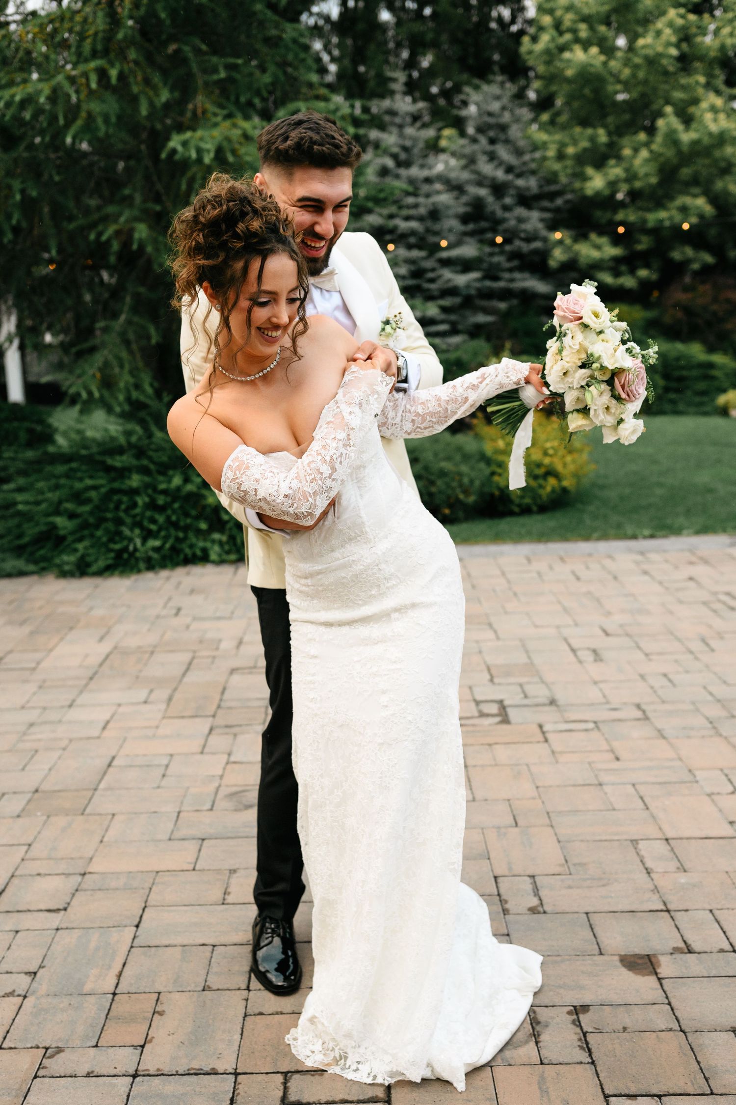 Joyful groom in white jacket embracing bride in lace wedding dress holding floral bouquet outdoors.