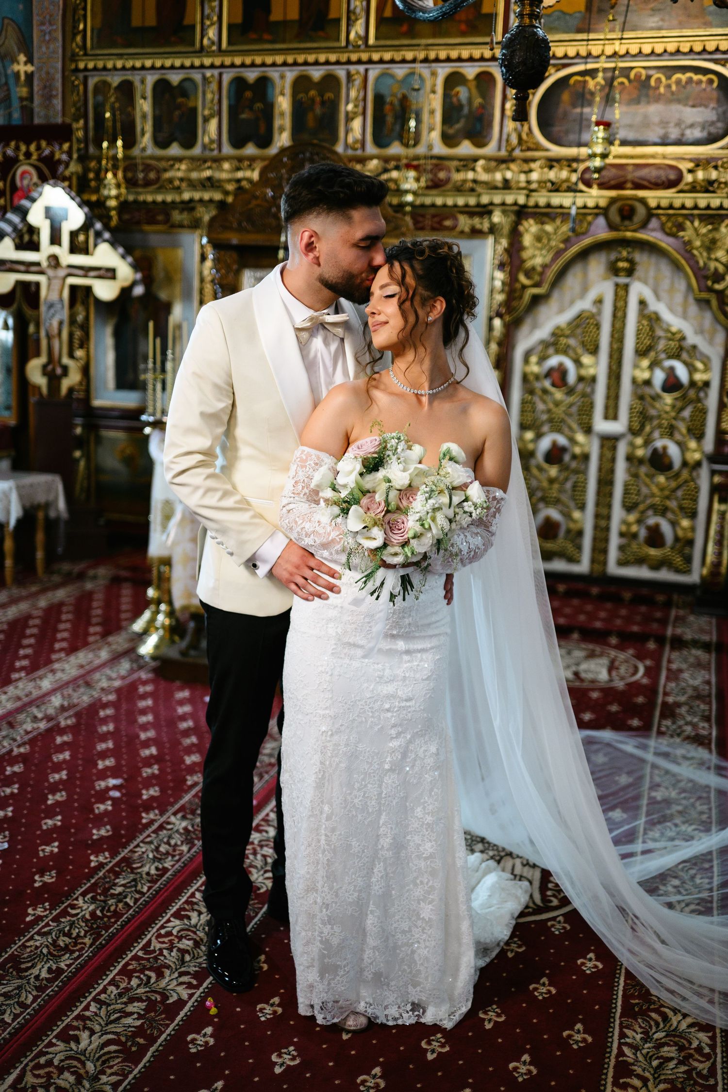 Bride and groom share a tender moment in an ornate Orthodox church, bride holding a white floral bouquet.