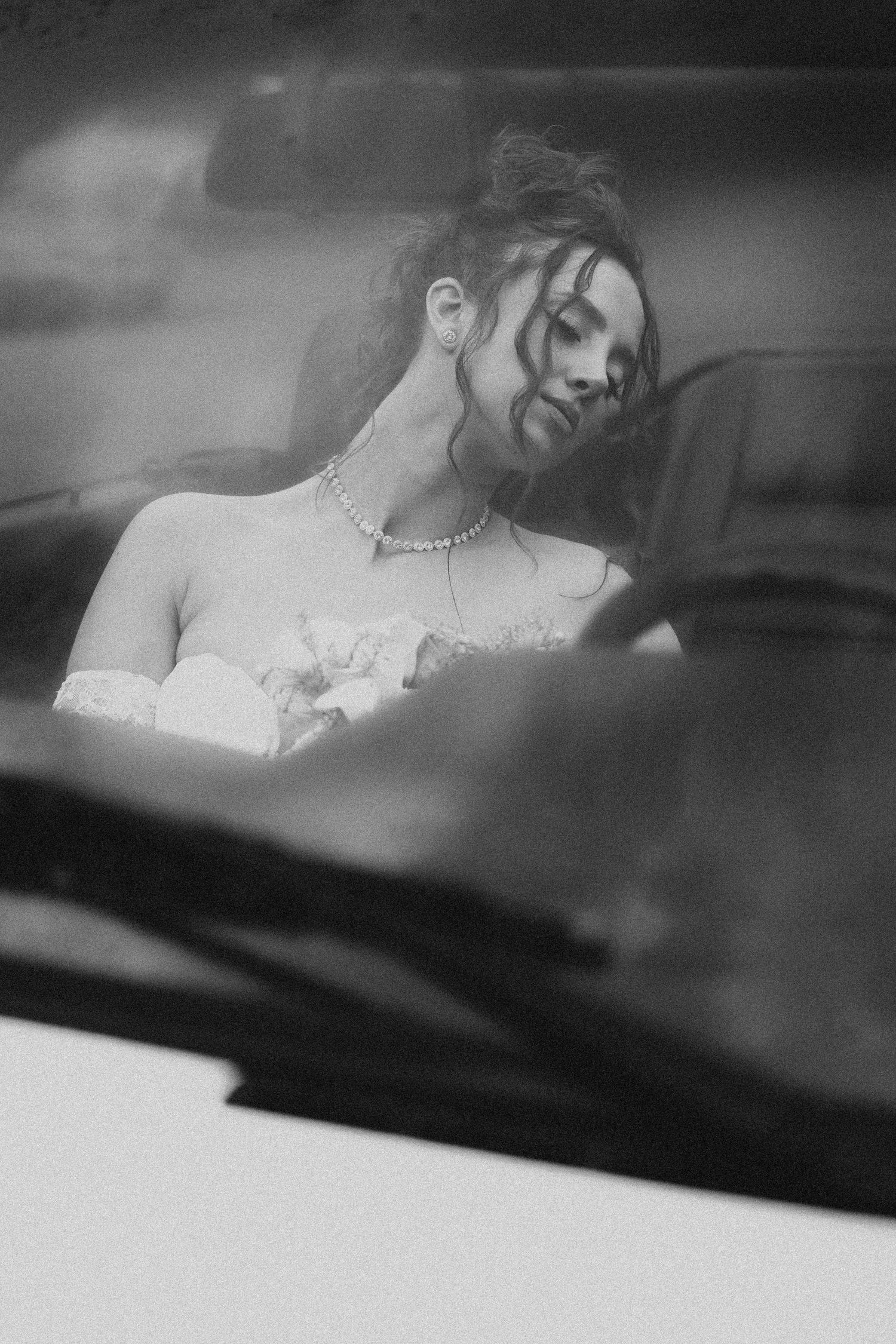 Black and white photo of a bride in a strapless gown and pearl necklace sitting in a car, looking down.
