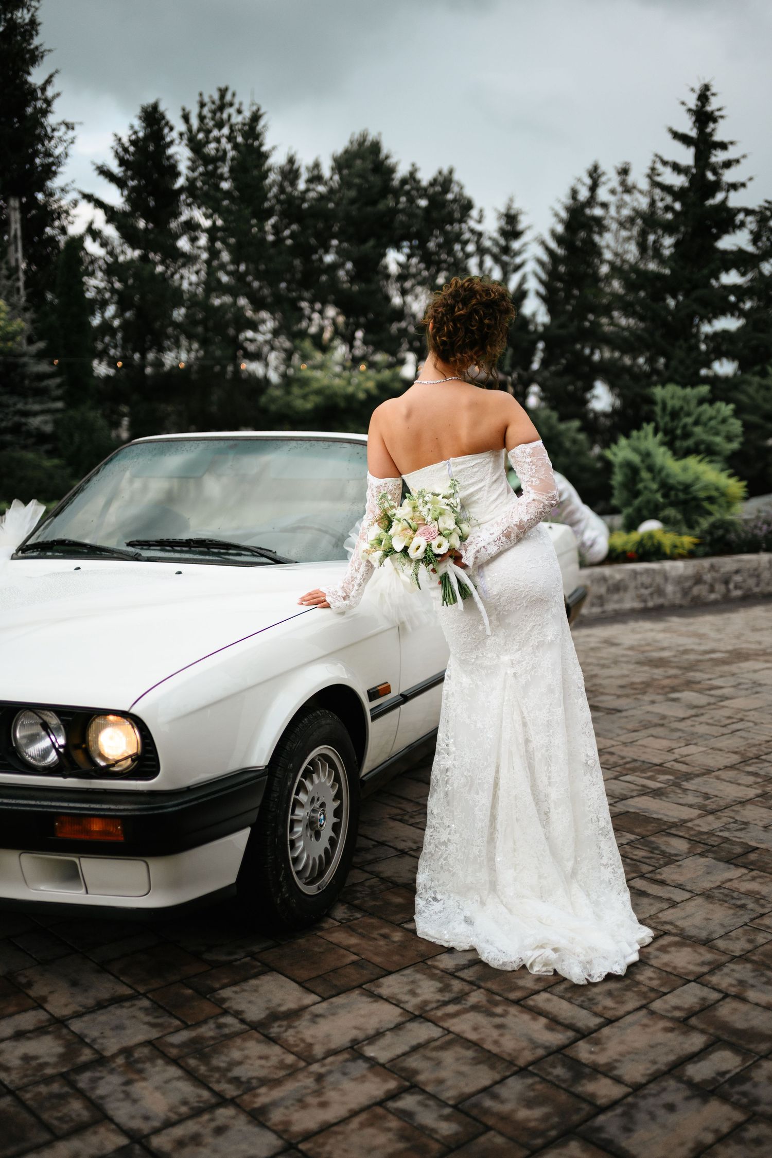 Bride in white lace gown holding bouquet stands beside a classic white BMW convertible with evergreen trees backdrop.