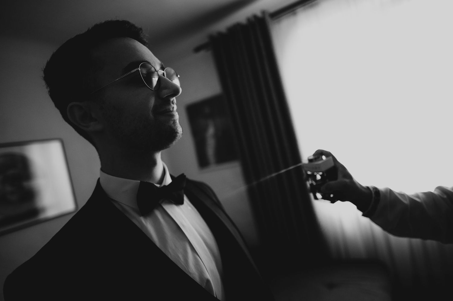 Black and white photo of a groom in a tuxedo and bow tie getting cologne sprayed before his wedding.
