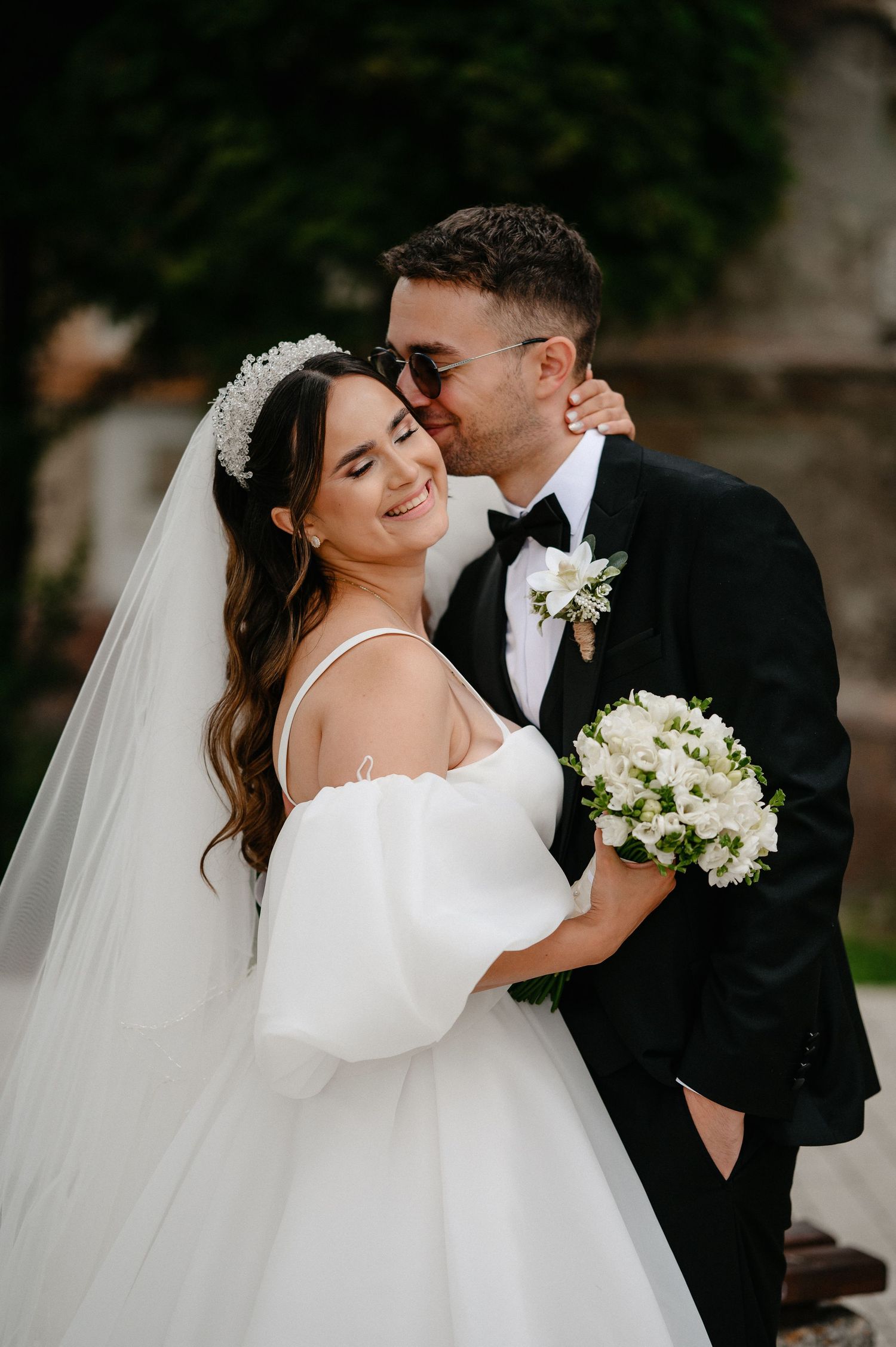 Groom kissing bride on cheek as she smiles holding a white bouquet on their wedding day.