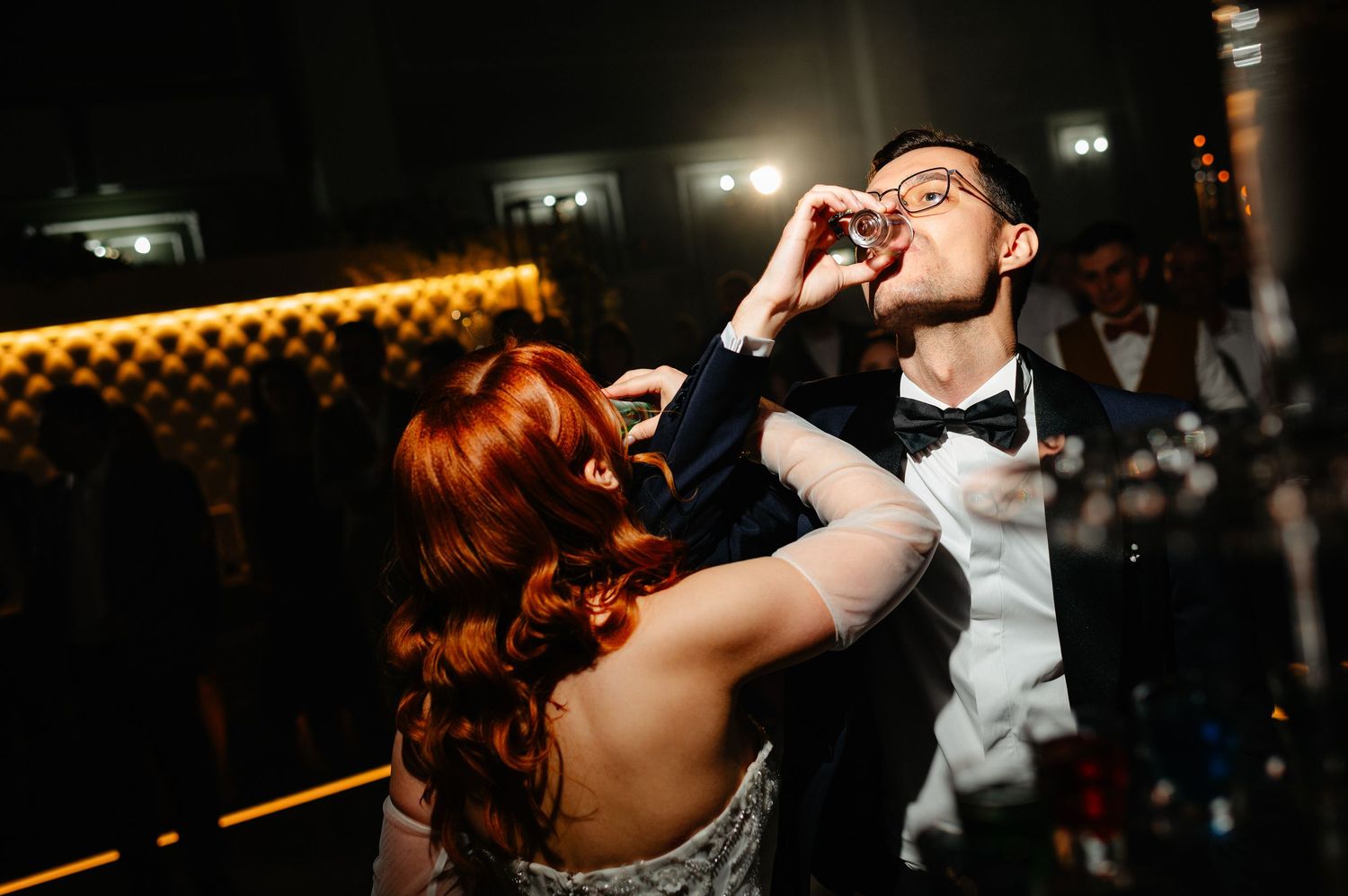 Bride with red hair feeds groom a shot glass drink during wedding reception dance floor celebration.