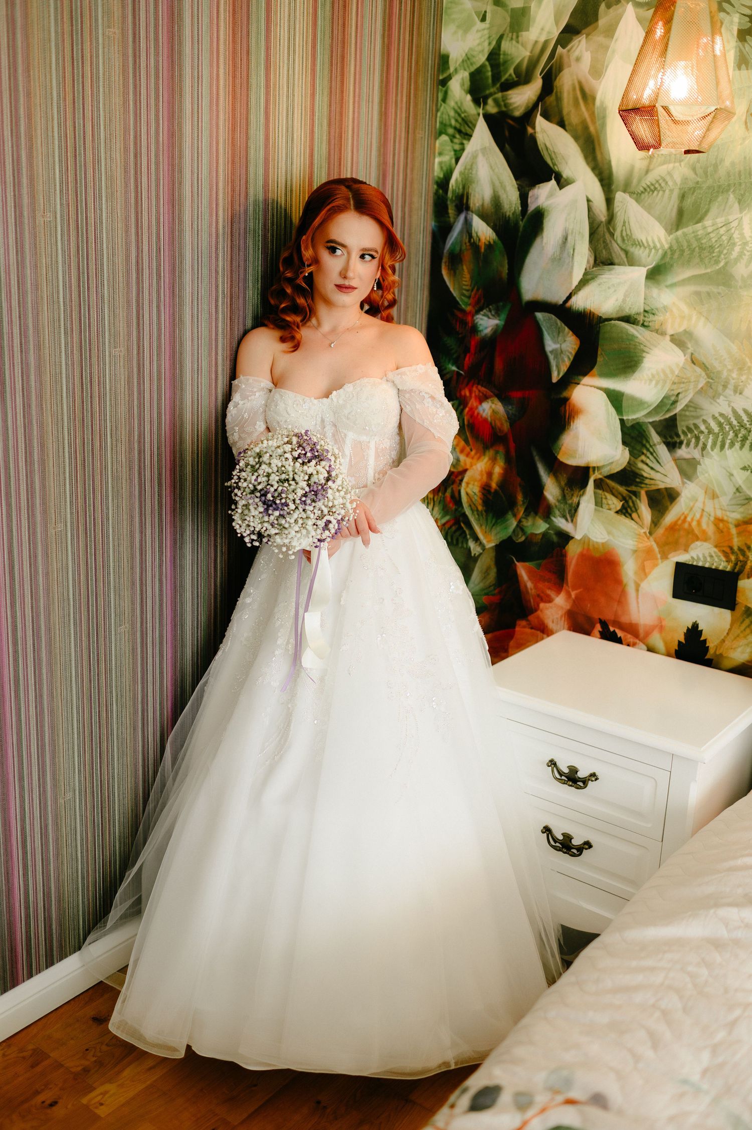 Red-haired bride in off-shoulder white ball gown holding bouquet, posing by floral wallpaper in elegant room.