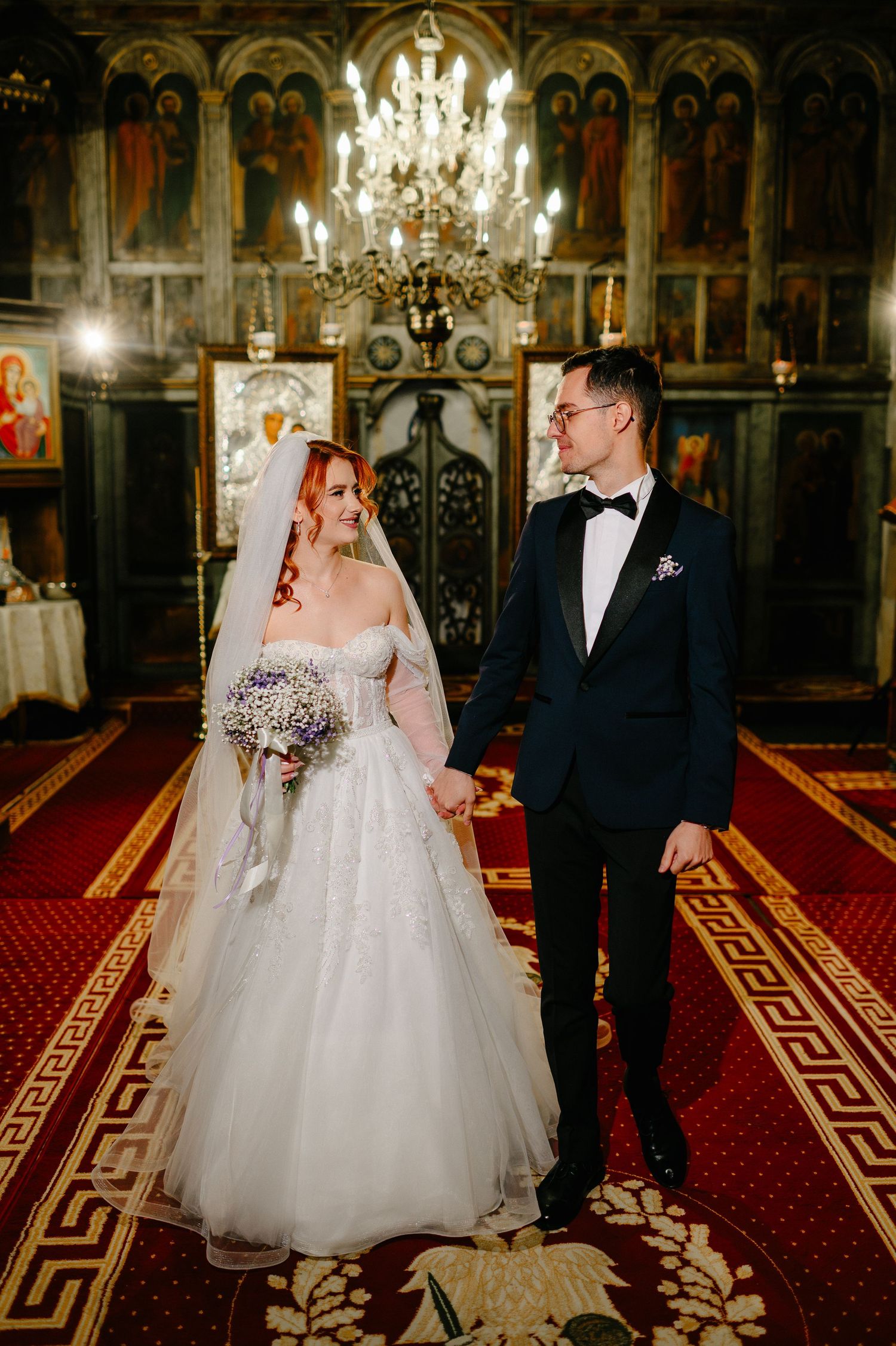 Bride and groom holding hands in an ornate Orthodox church, bride in white gown with veil, groom in black tuxedo.