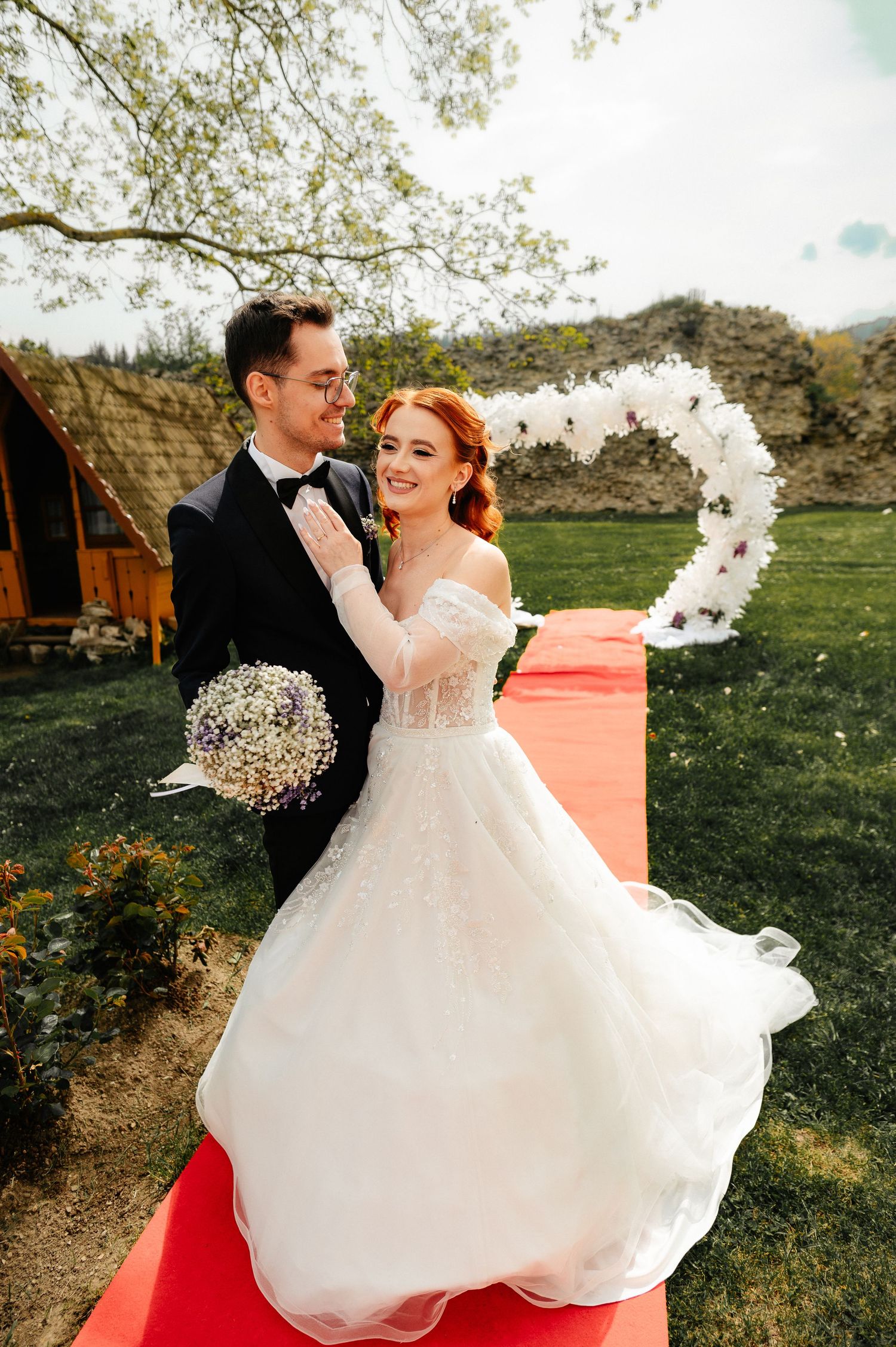 Bride in white gown holds bouquet beside groom in tuxedo on red carpet at outdoor wedding ceremony.