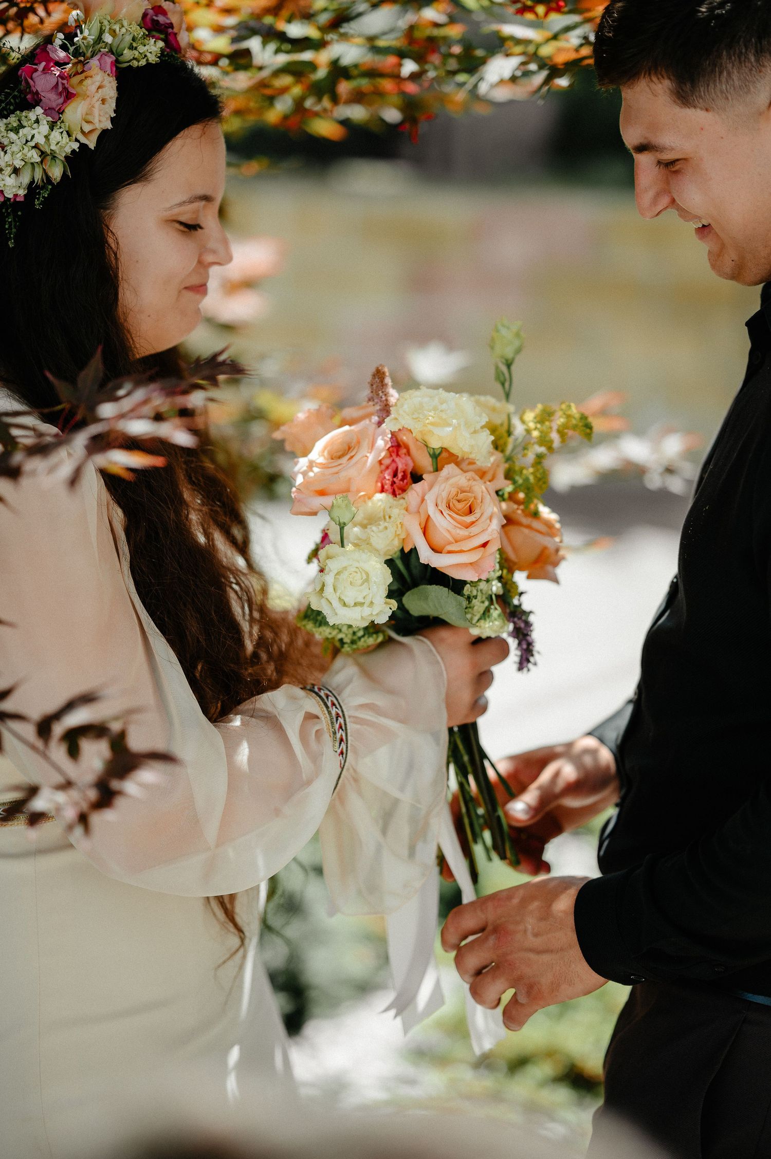 Bride in white dress with floral crown exchanges rings with groom, holding peach and white rose bouquet outdoors.