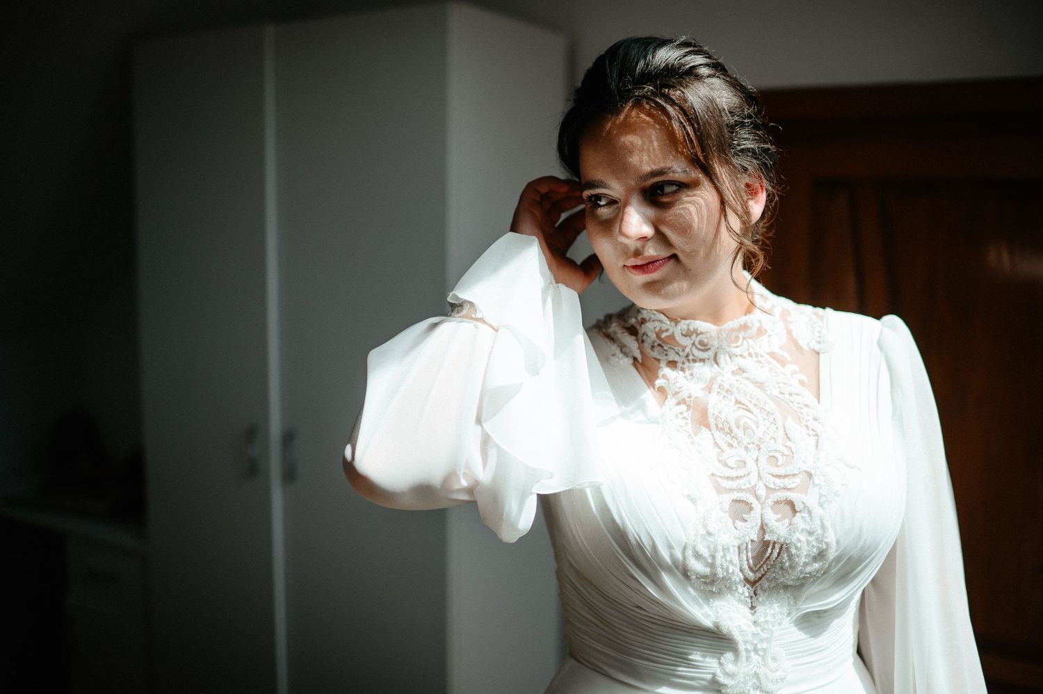Bride in elegant white lace wedding gown adjusting her earring while getting ready indoors.