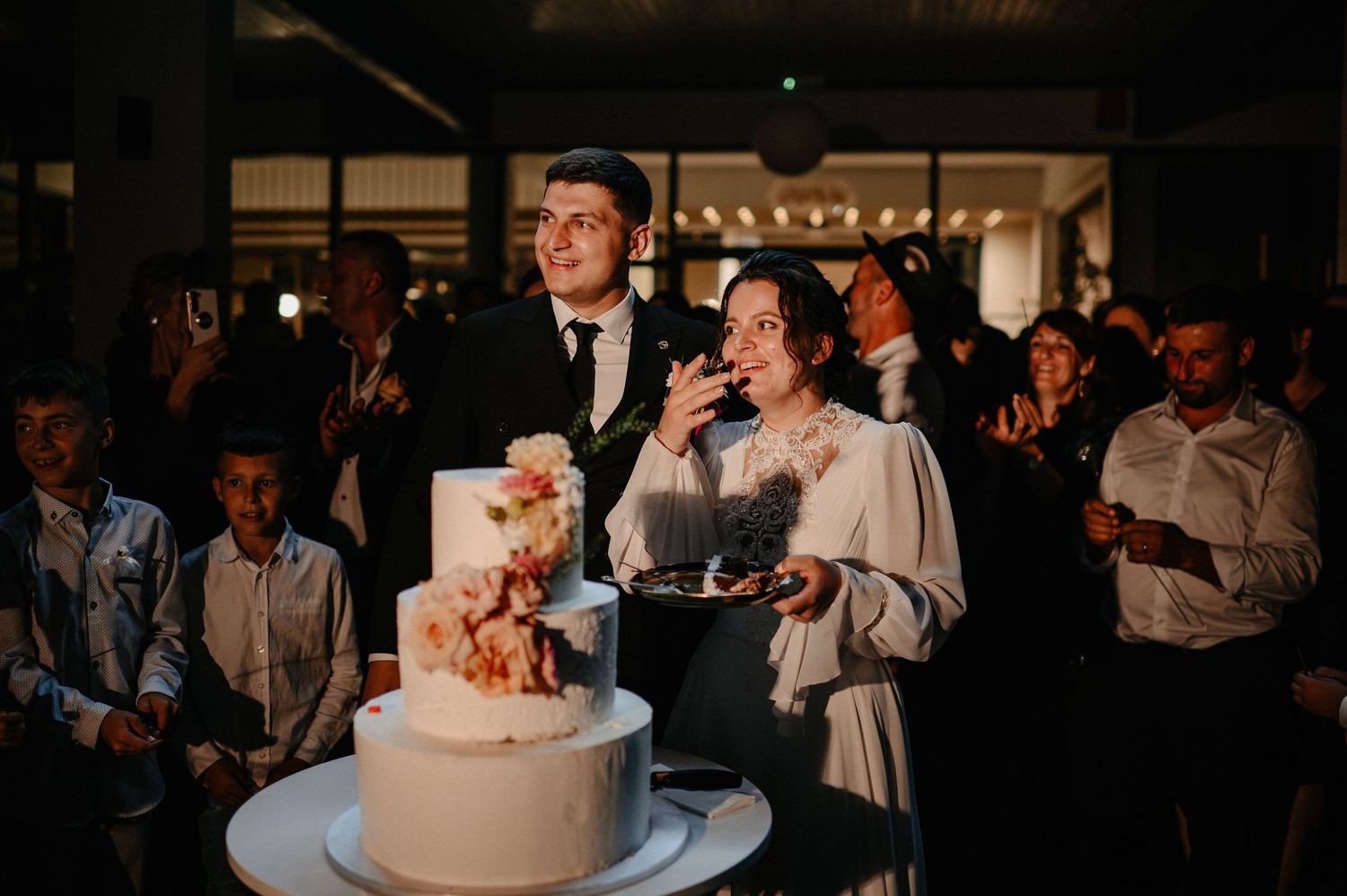 Newlyweds cut elegant three-tier white wedding cake surrounded by guests at reception.