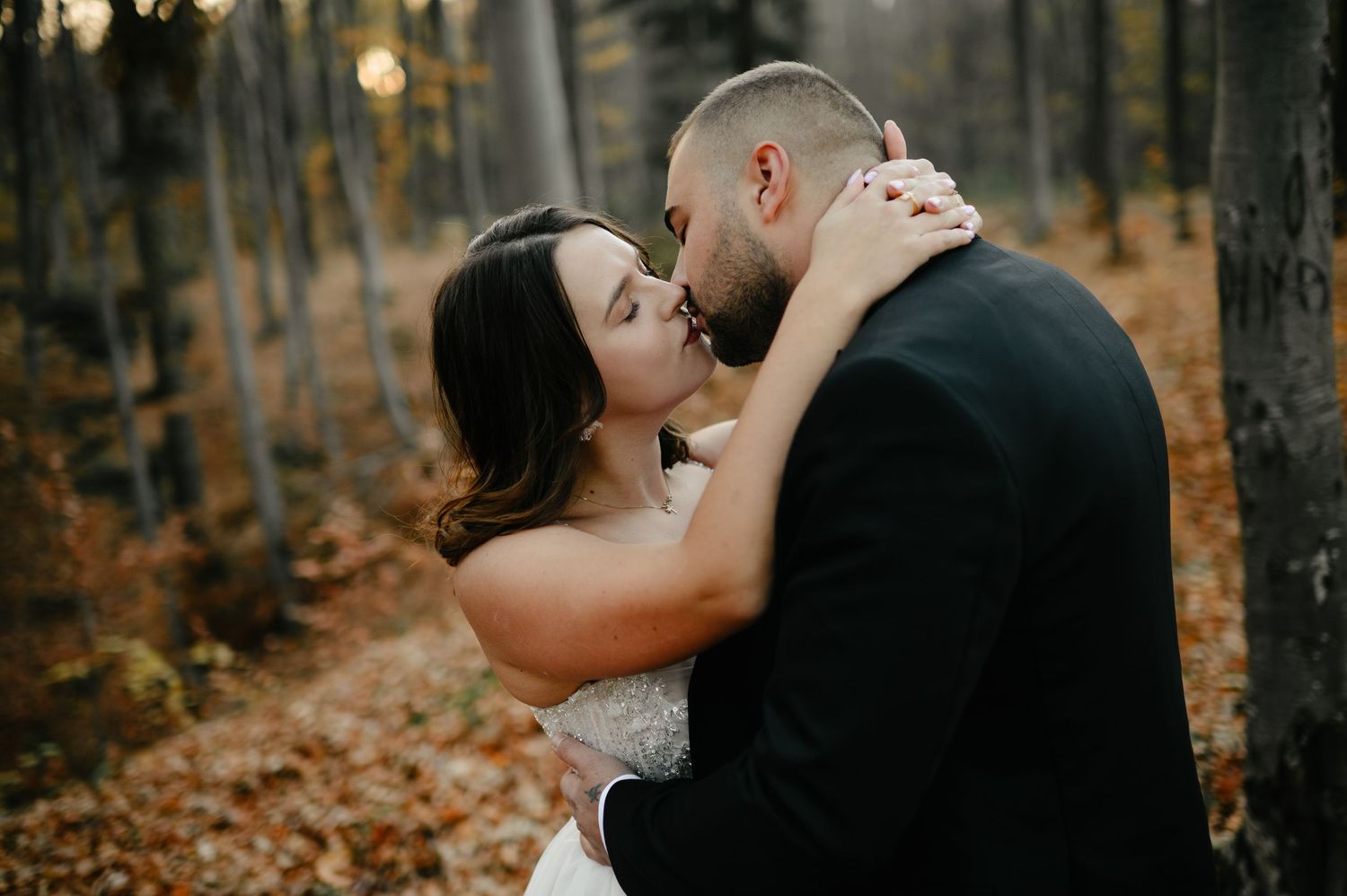 A bride and groom share a romantic kiss surrounded by autumn foliage in a serene forest setting.