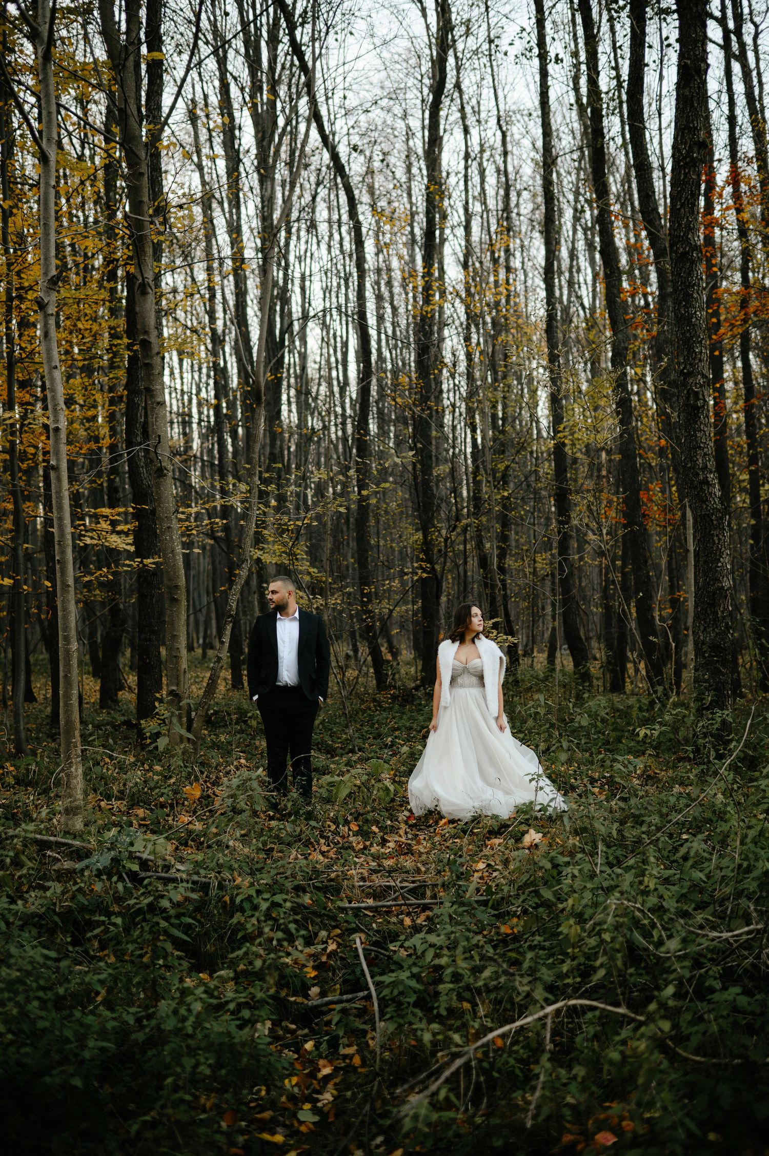 Bride in white gown and groom in black suit stand apart in an autumn forest with colorful foliage.