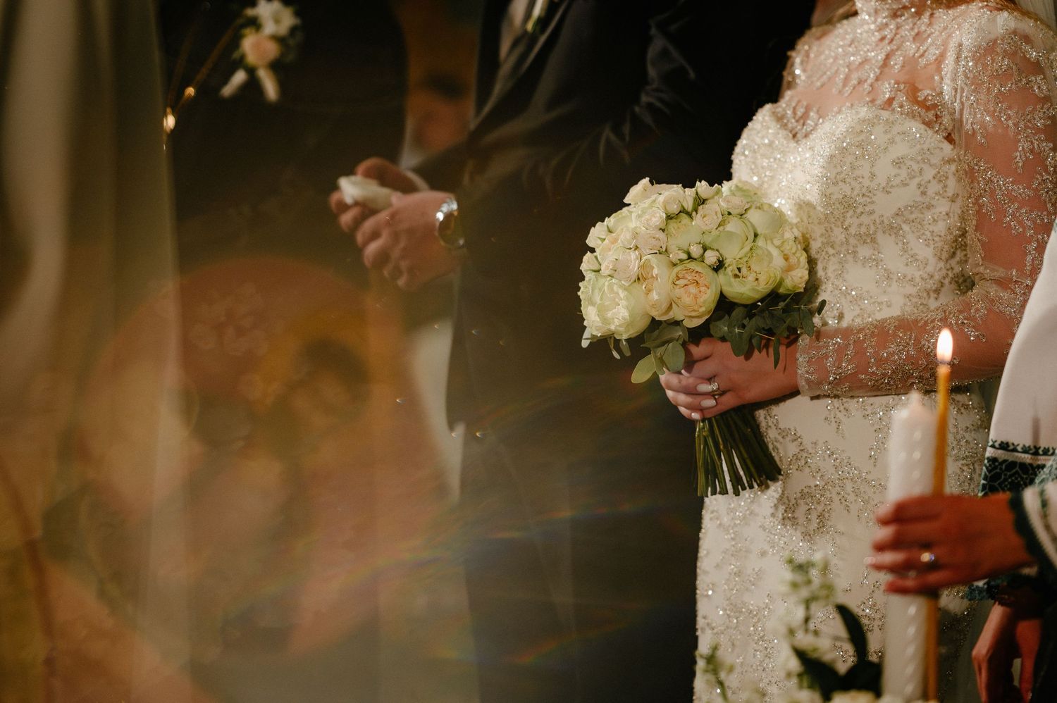 Bride holding white rose bouquet beside groom at wedding ceremony with candlelight.