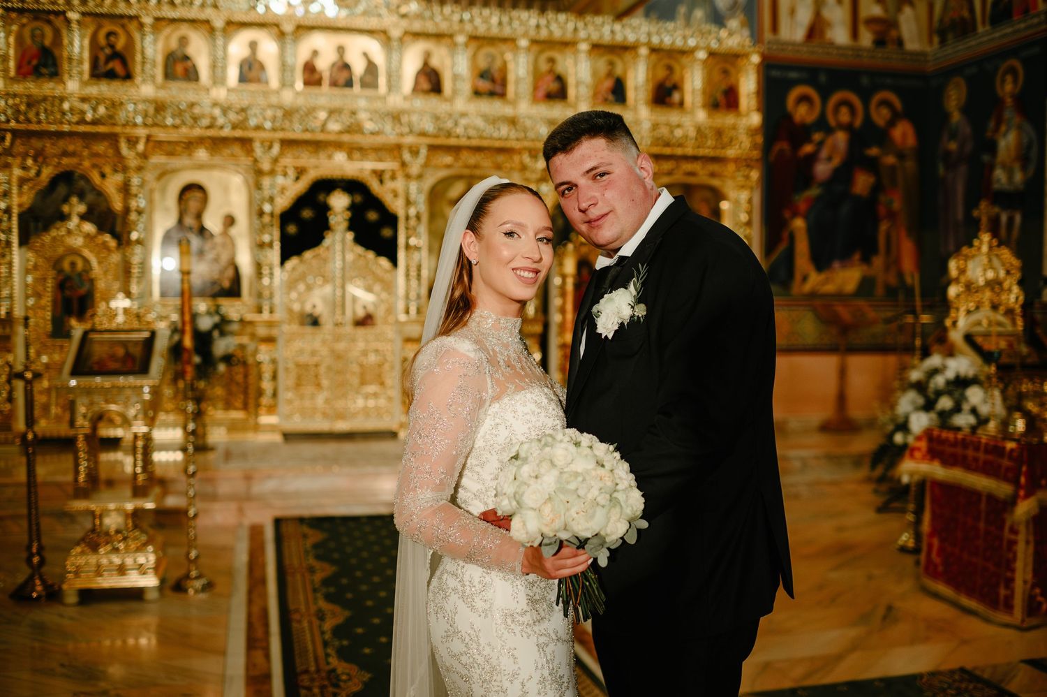 Newlywed couple poses together in front of a golden Orthodox church iconostasis after their ceremony.