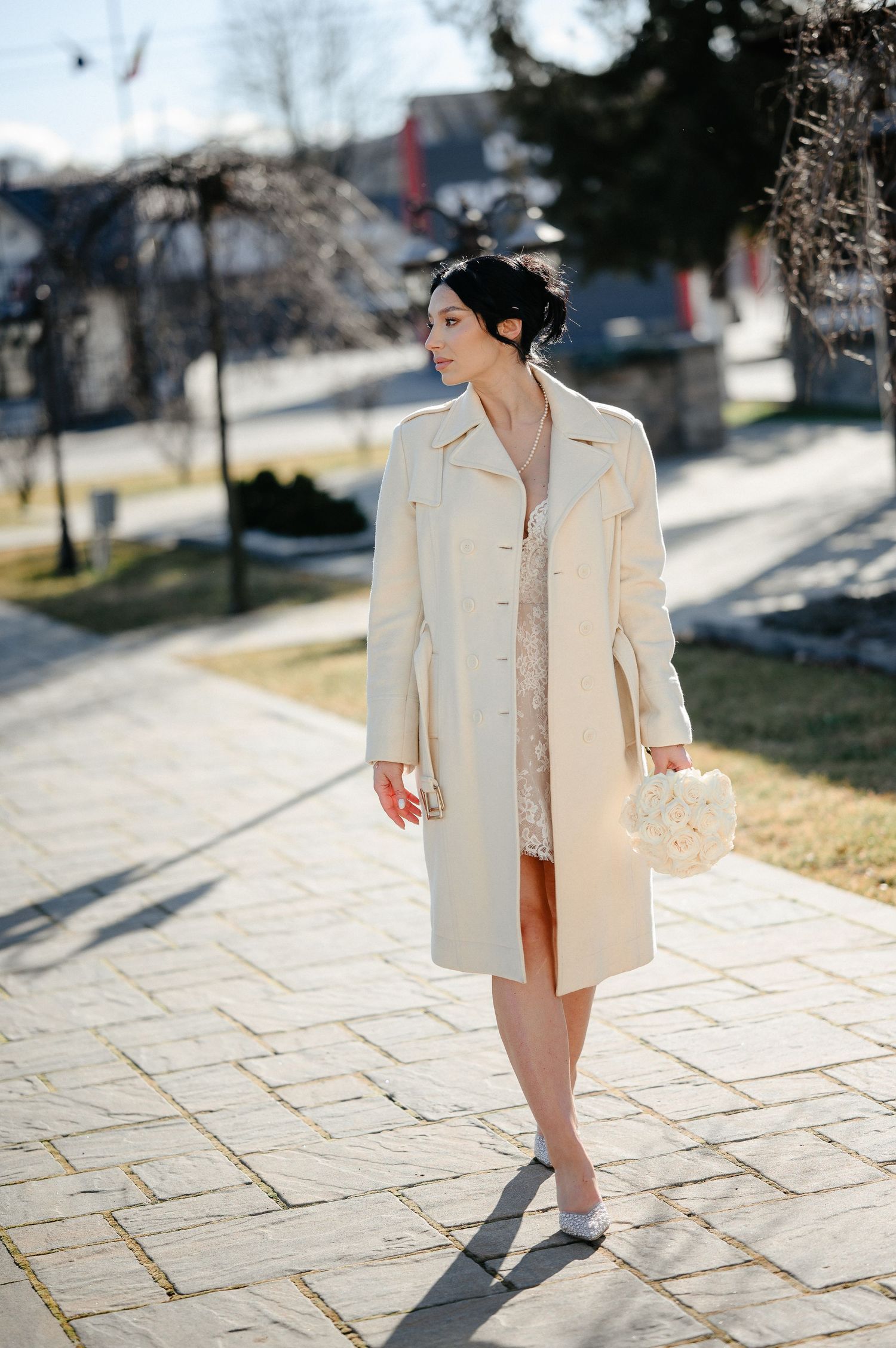 Elegant bride in cream coat holding bouquet walks sunny path.