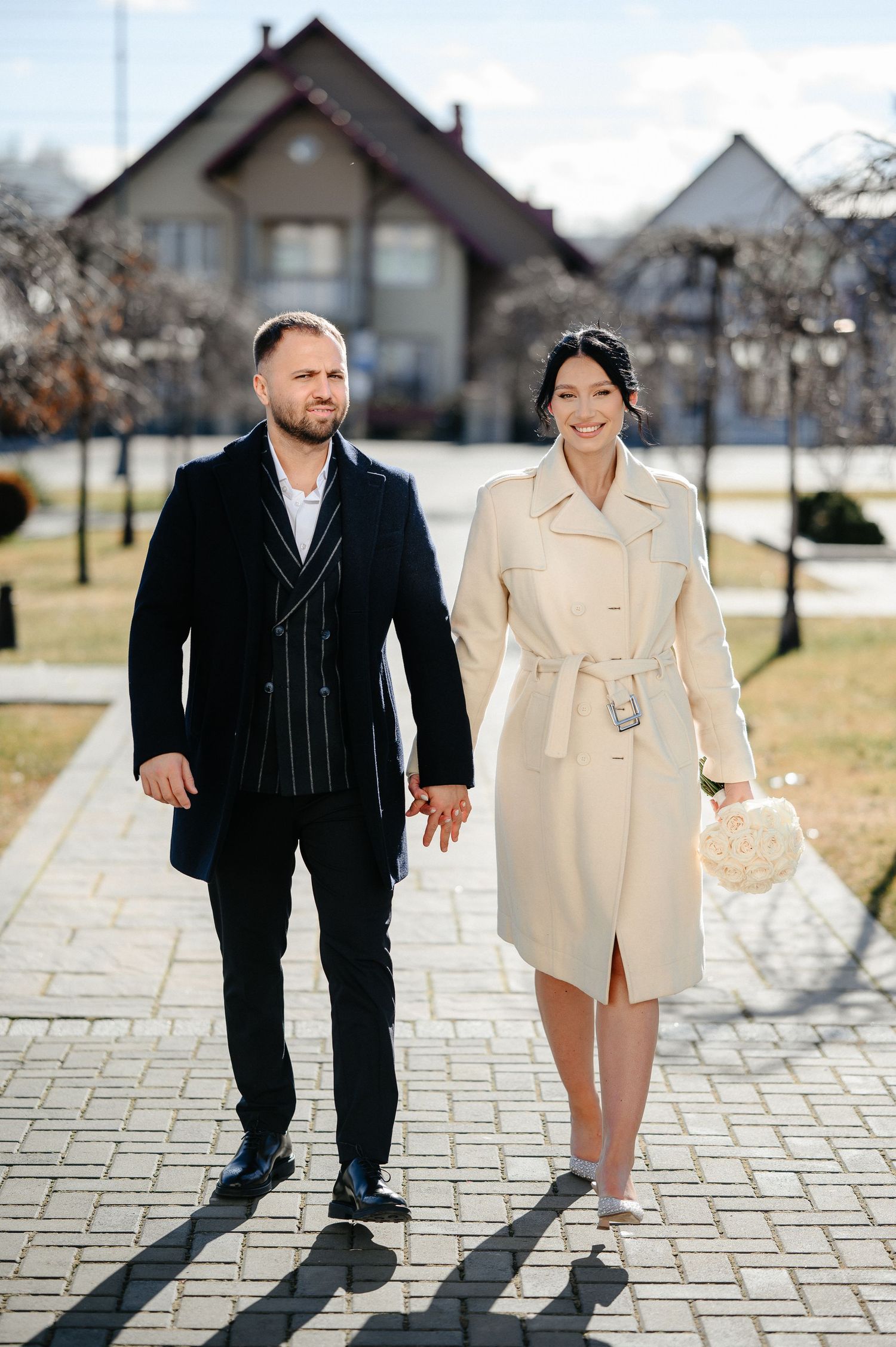 Elegant couple holding hands walking on a brick path outdoors, man in dark suit, woman in cream trench coat.