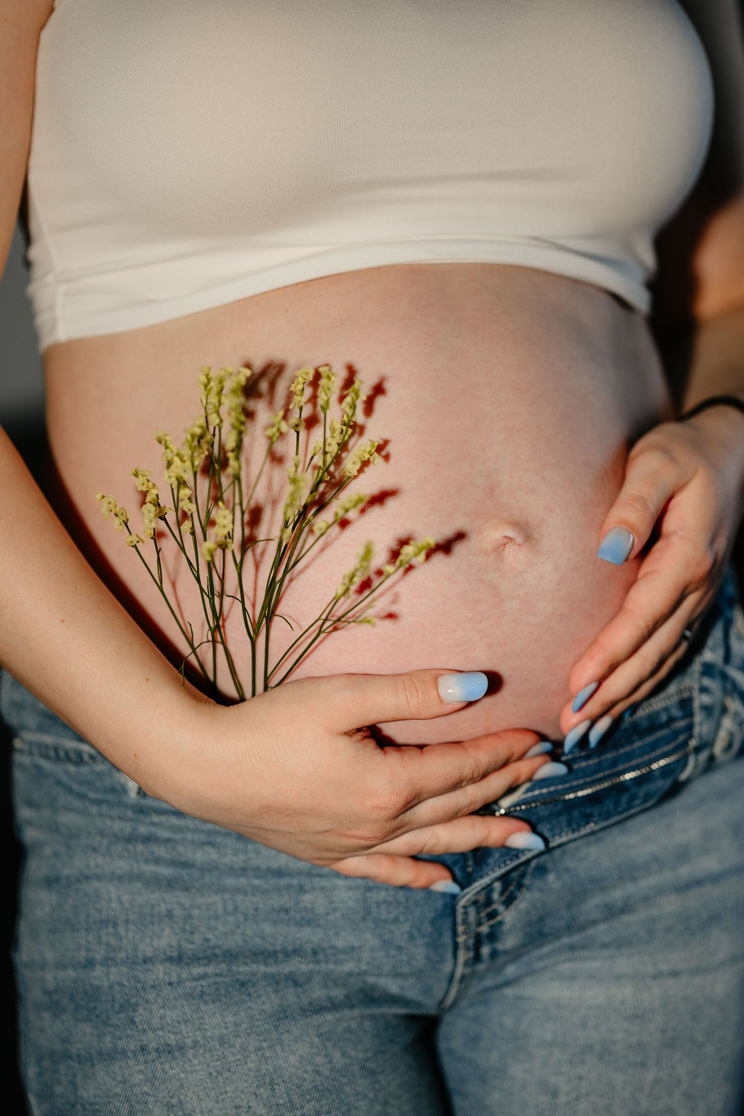 Pregnant woman in white crop top and jeans cradling baby bump with delicate wildflowers resting on her belly.