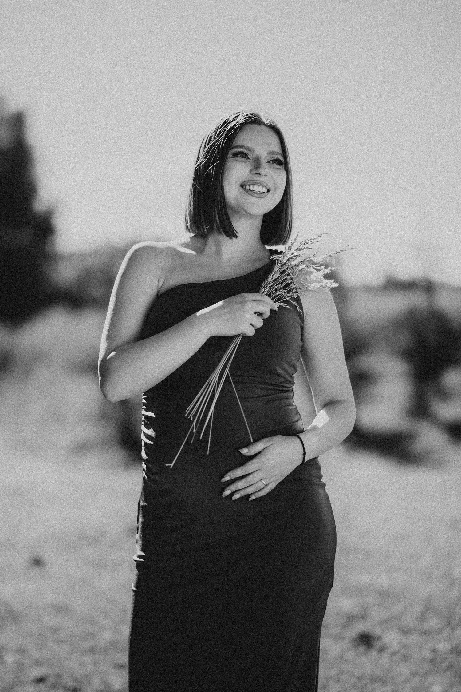 Young woman in black one-shoulder dress holds wildflowers in a black and white outdoor portrait.