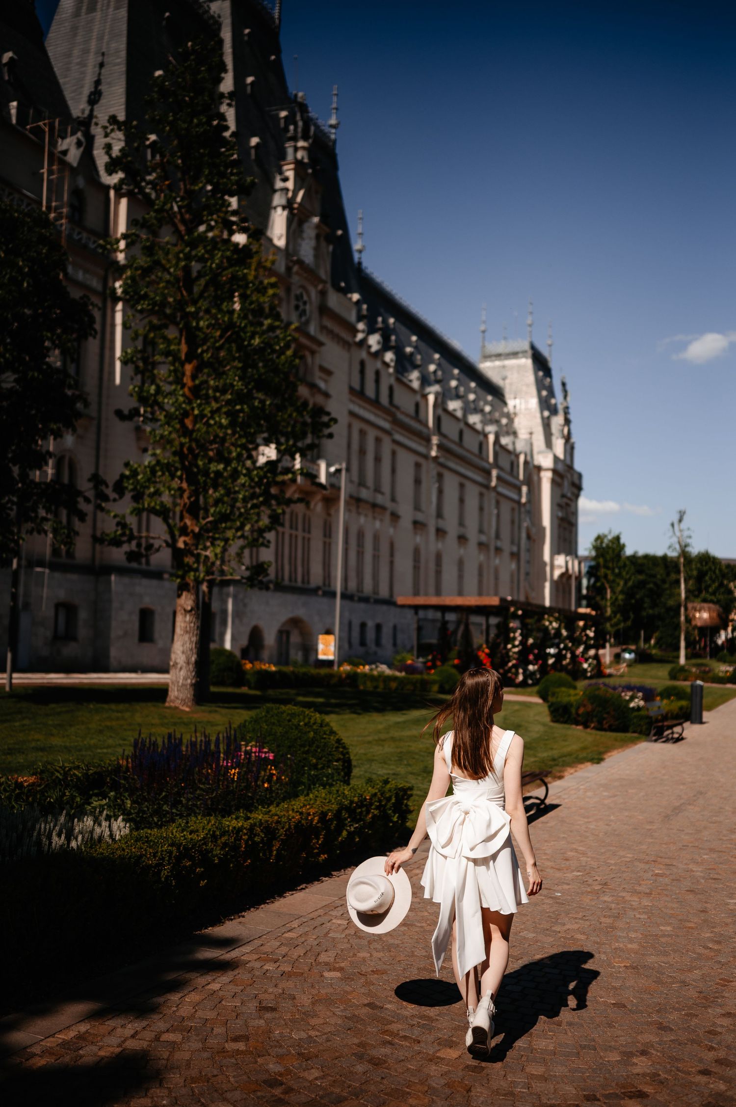 Woman in white dress carrying a hat walking along a path beside a grand historic building.