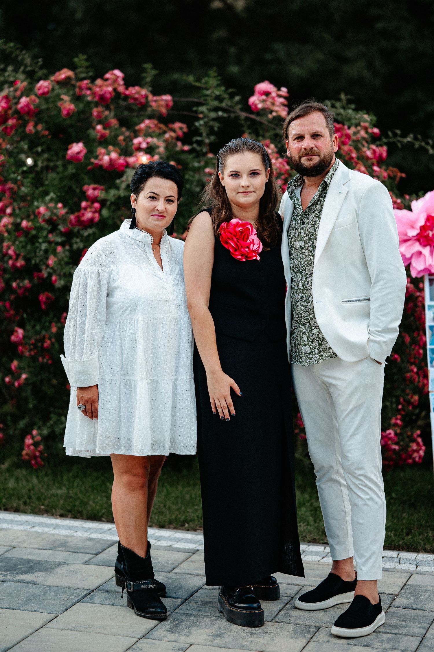 Family of three posing formally in front of blooming pink roses, woman in black dress with red flower.