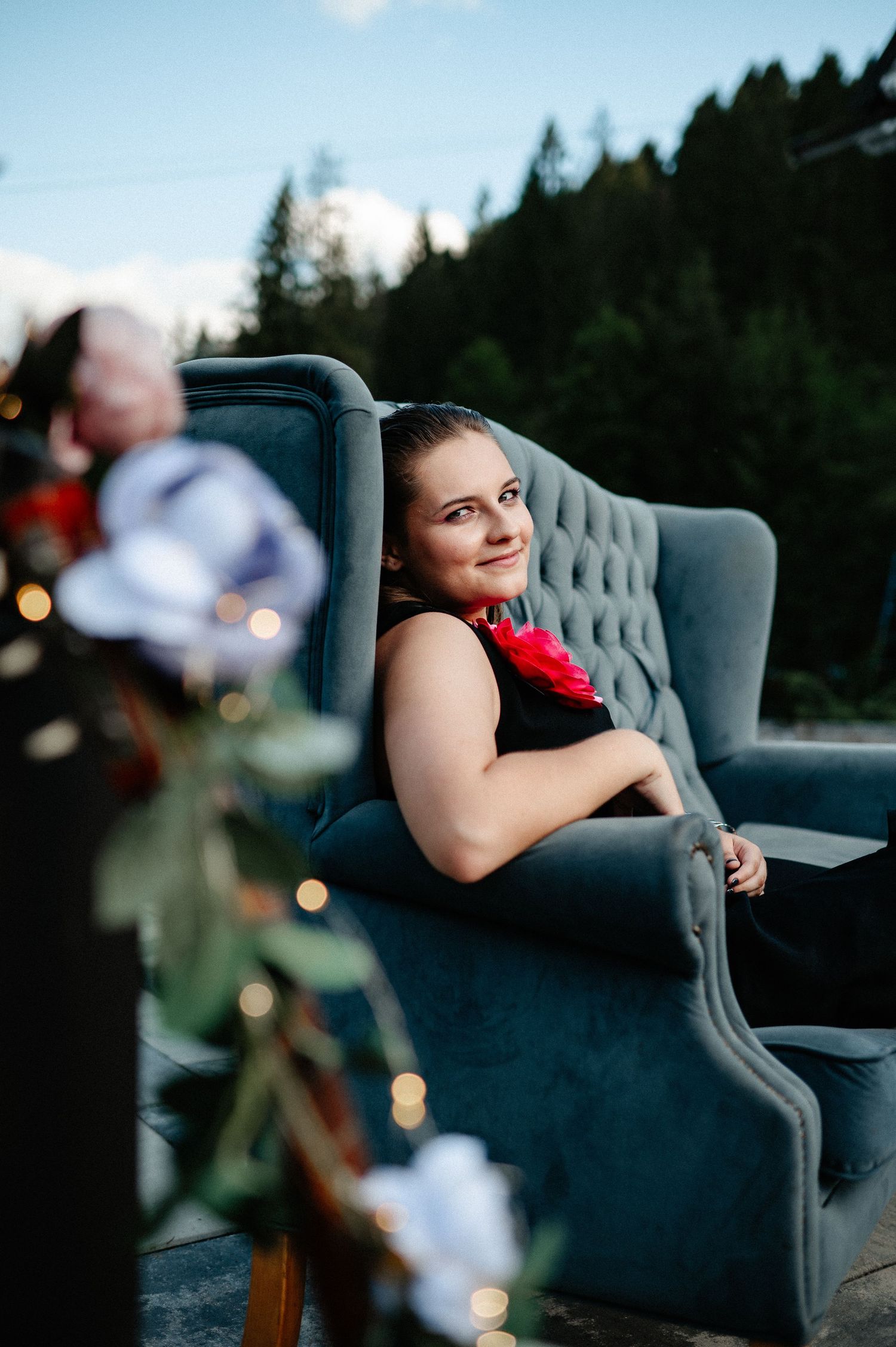 Young woman in black dress with red flower relaxing in a tufted gray velvet armchair outdoors.