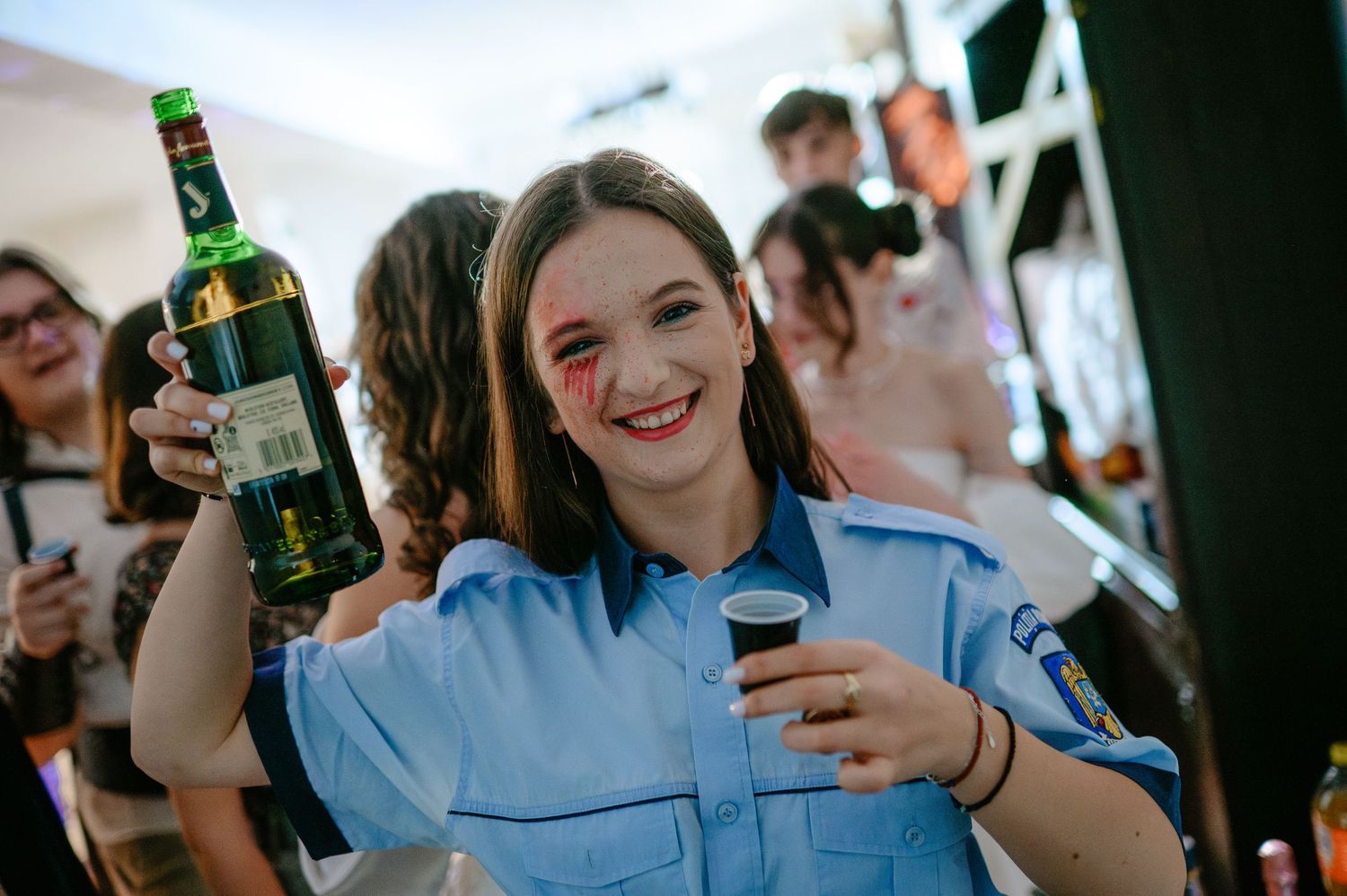 A smiling woman in a police costume holds a green bottle and glass at a Halloween party.