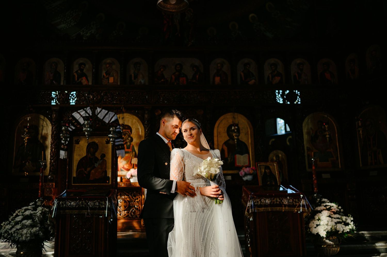 Wedding couple standing together in front of ornate Orthodox church iconostasis with golden religious icons.