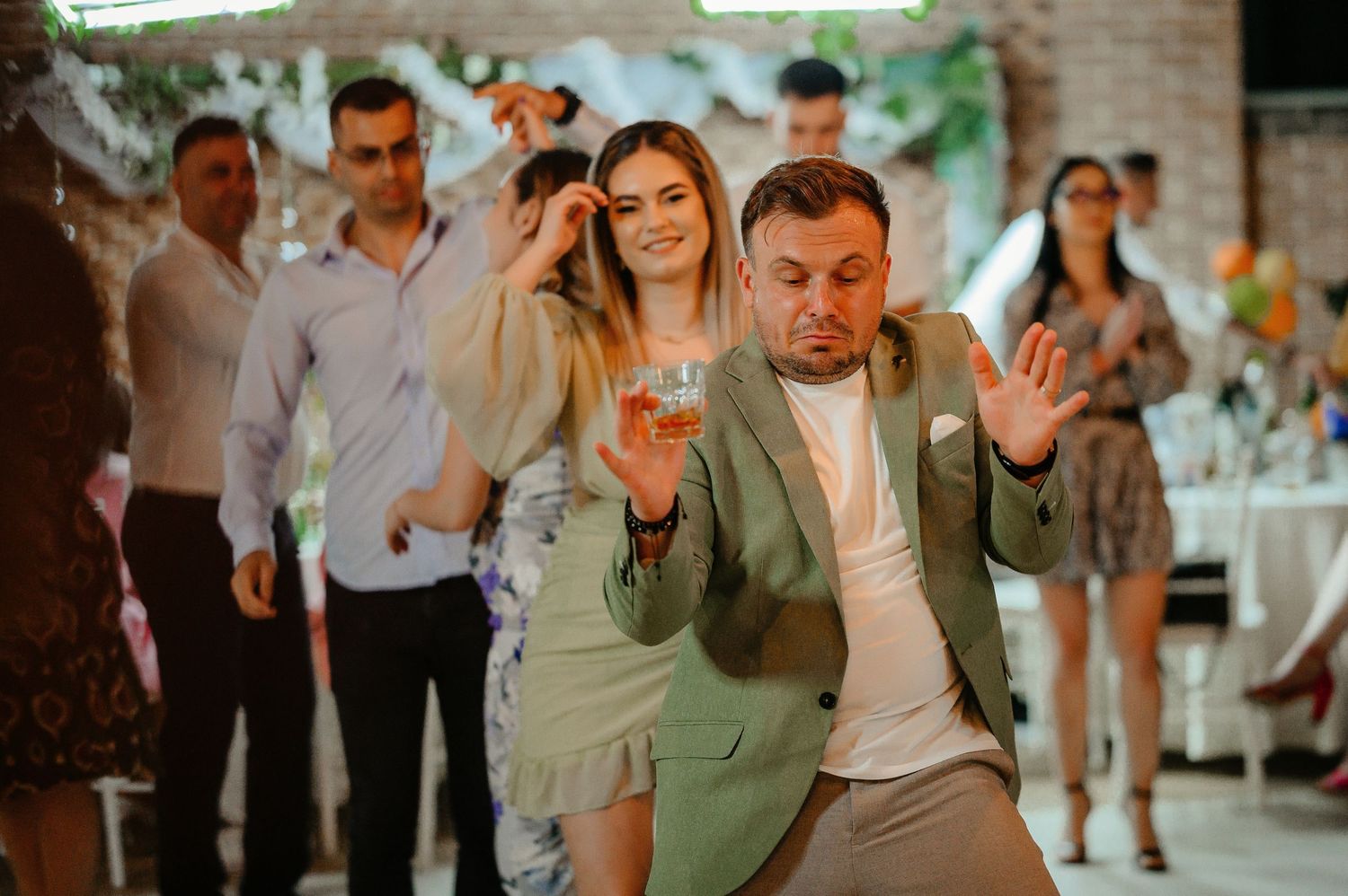 A man in a green blazer dances energetically at a celebration while a smiling woman watches behind him.