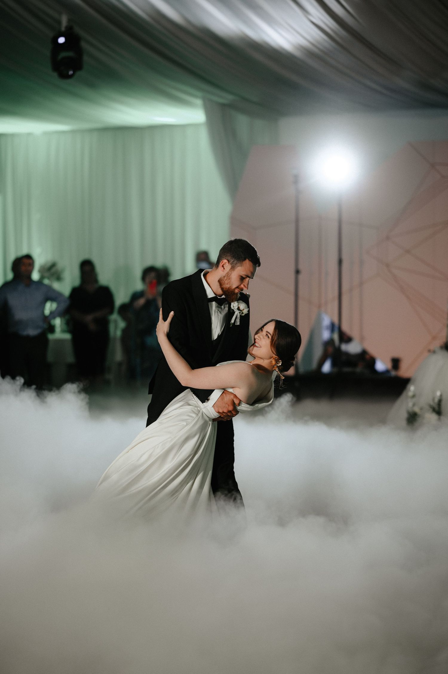 Bride and groom share romantic first dance surrounded by dramatic fog on elegant wedding reception dance floor.