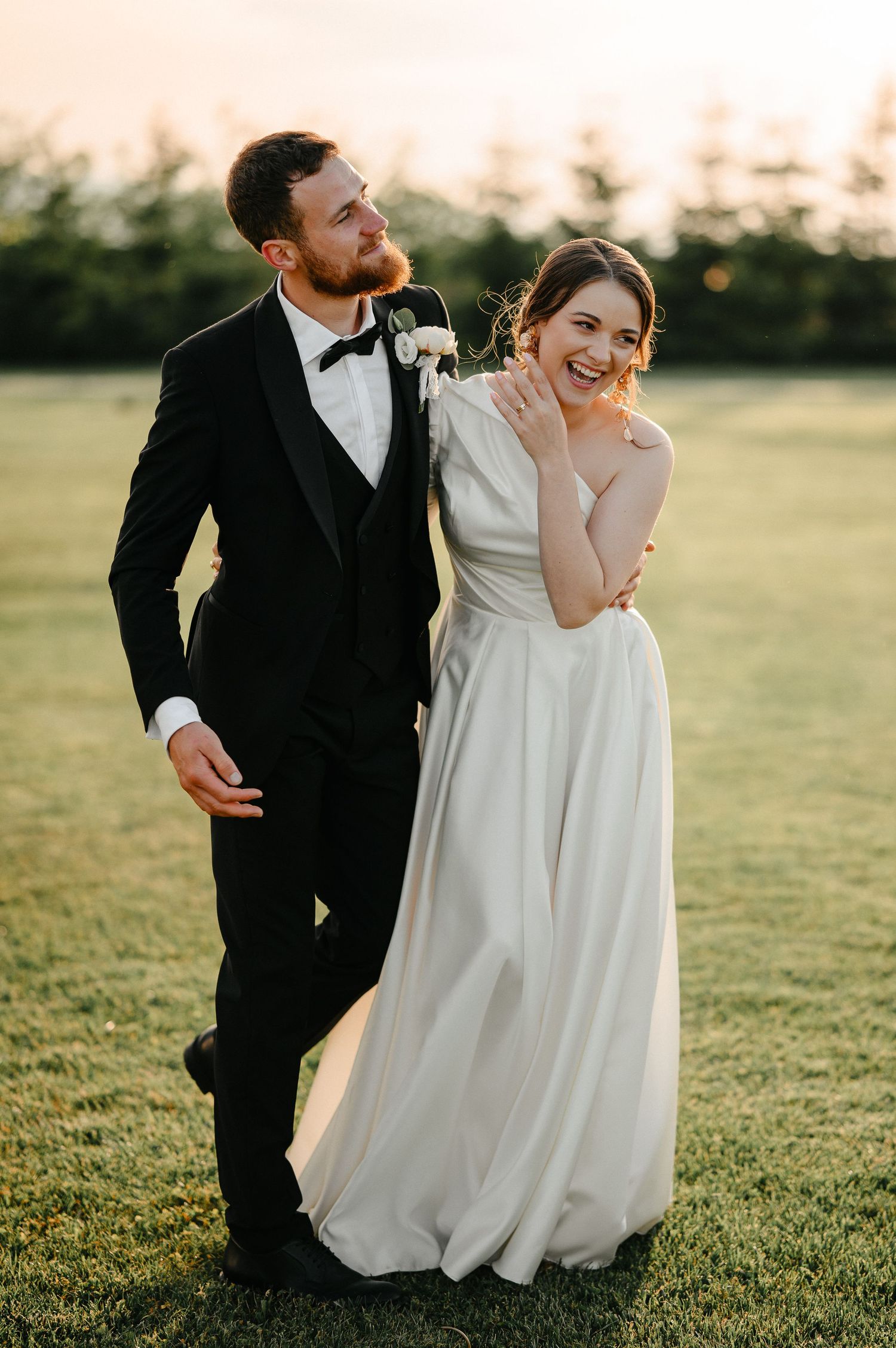 Bride and groom laughing together on green lawn at golden hour sunset wedding.