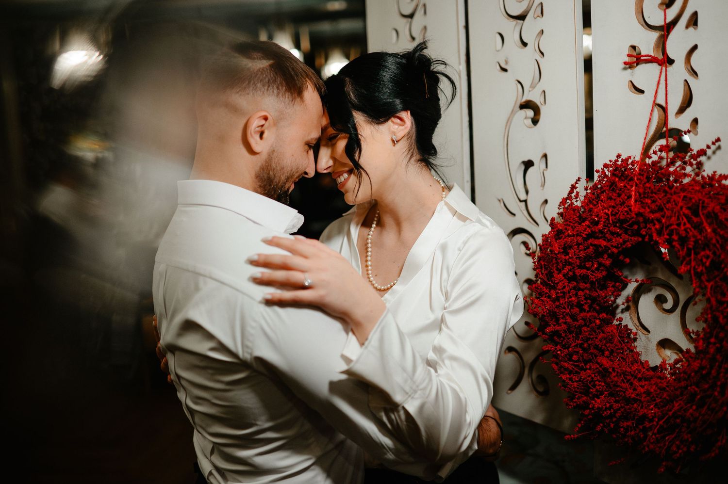 Couple in white attire sharing romantic moment near red floral wreath decoration.