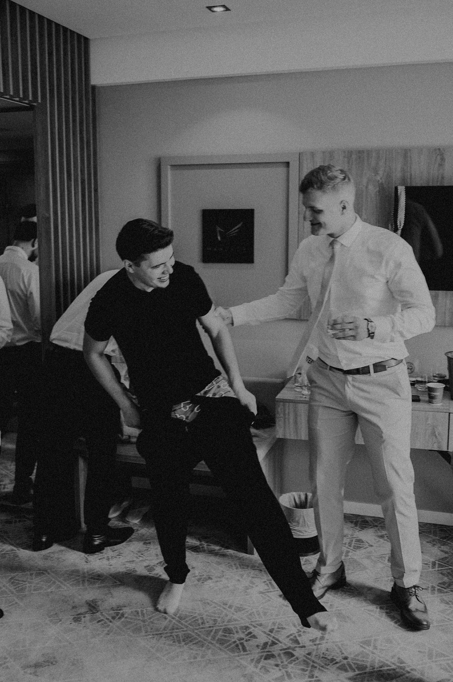 Black and white photo of two young men laughing and dancing together in a hotel room before a wedding.