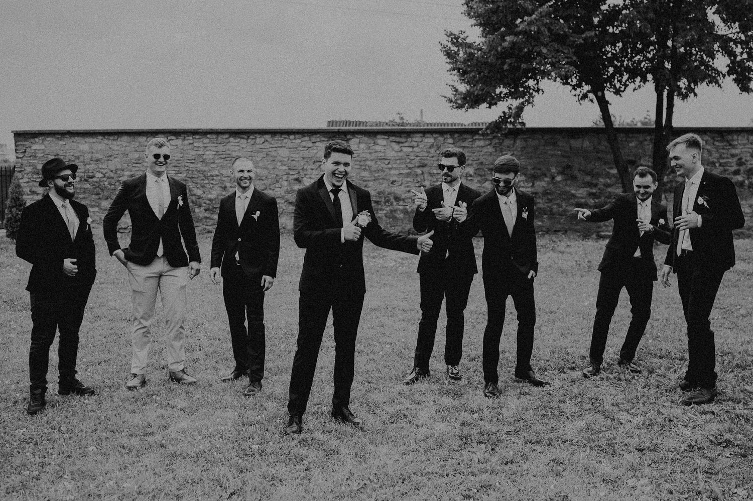 Black and white photo of groom and groomsmen posing casually in dark suits on a grassy field.