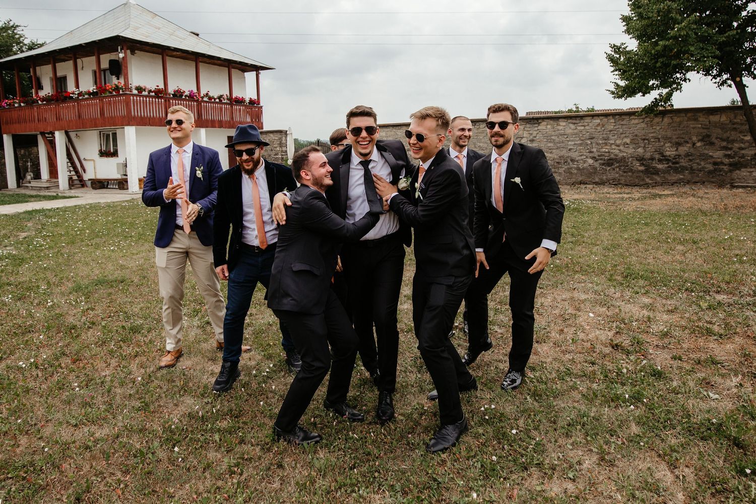 Energetic groomsmen in dark suits playfully surrounding the groom outdoors during wedding day celebrations.