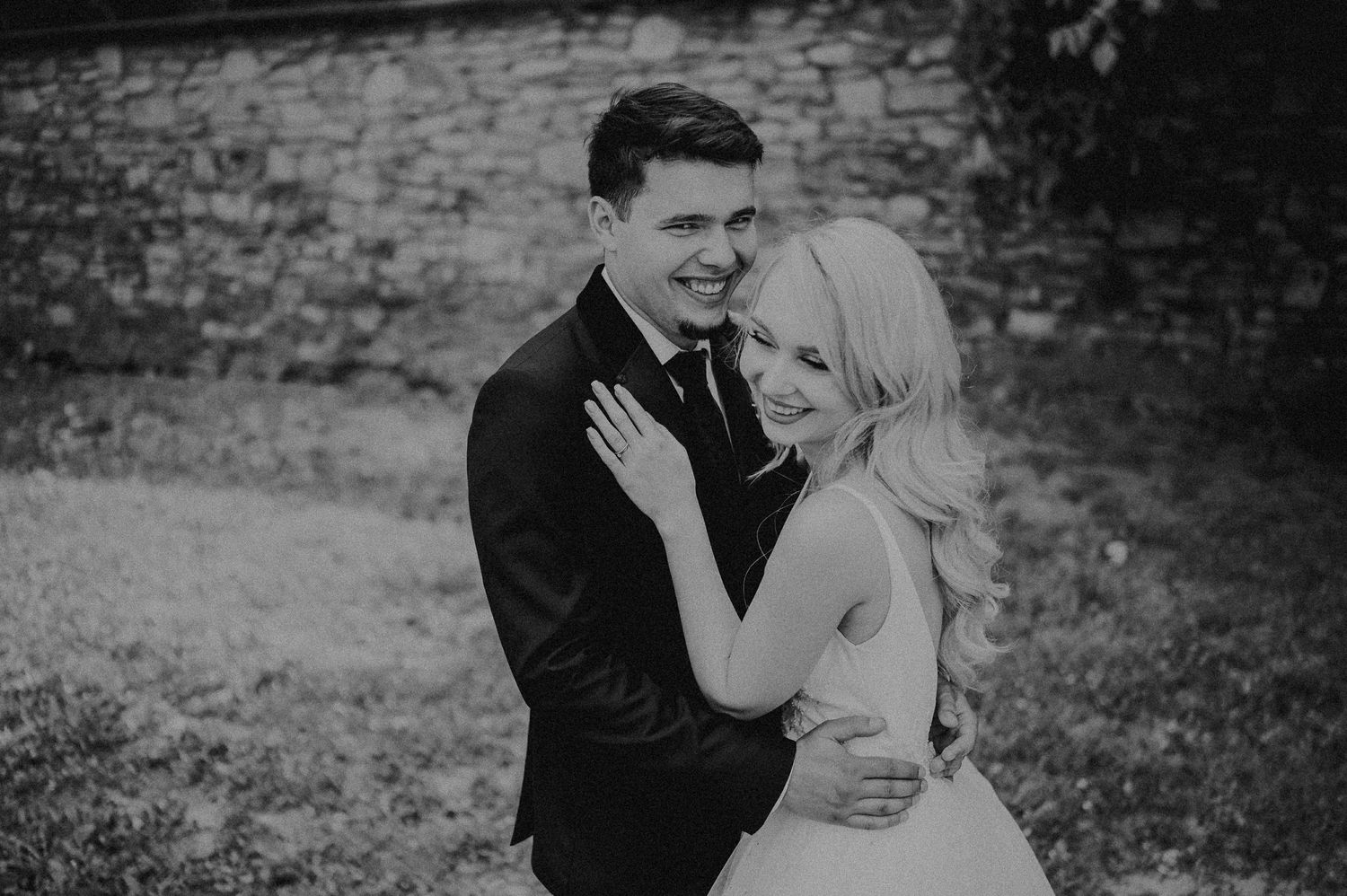 Black and white photo of smiling bride and groom embracing outdoors against a stone wall.